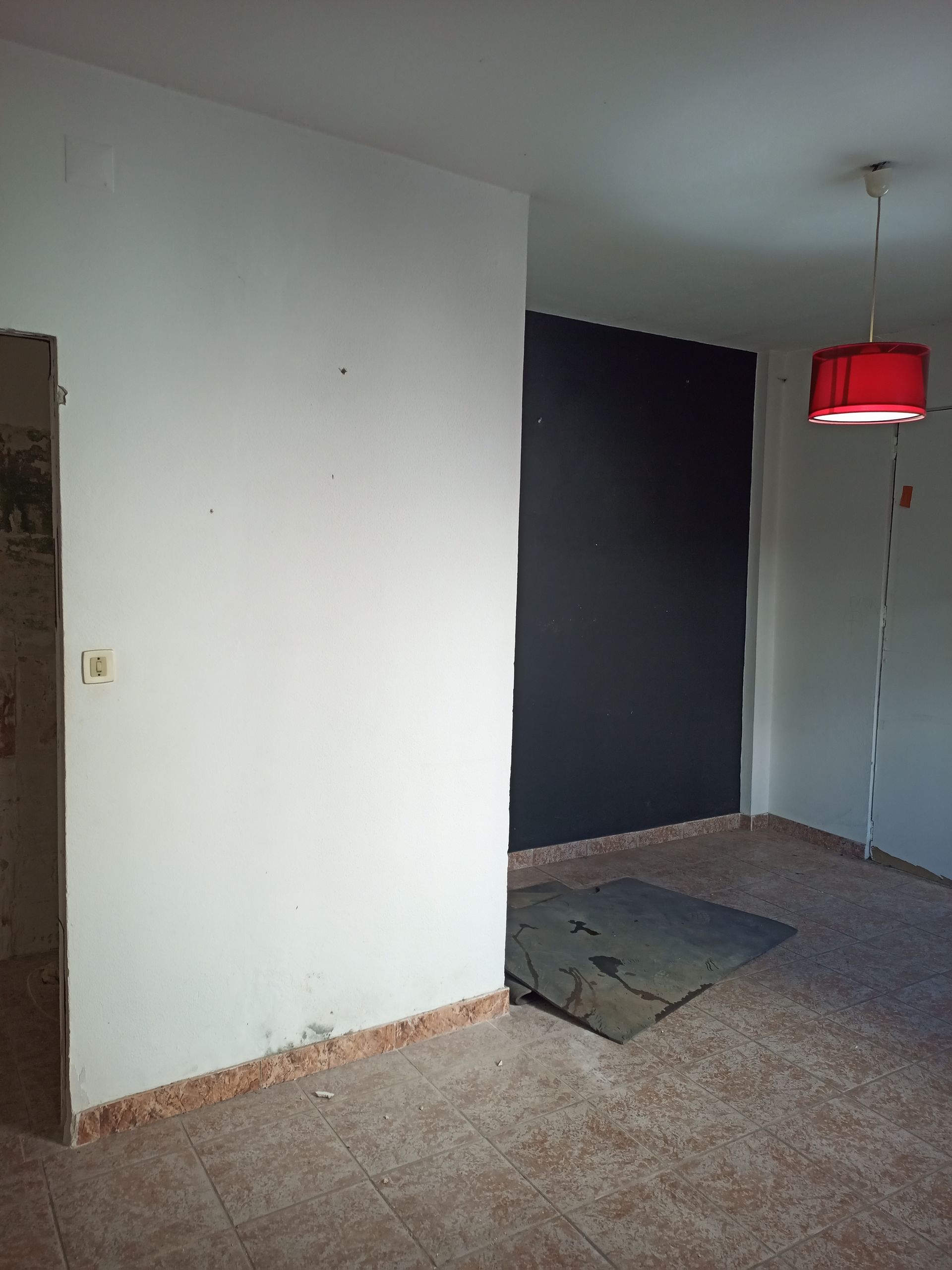 Quarto vazio com paredes brancas e pretas, abajur vermelho. Piso de azulejo marrom, entulho.