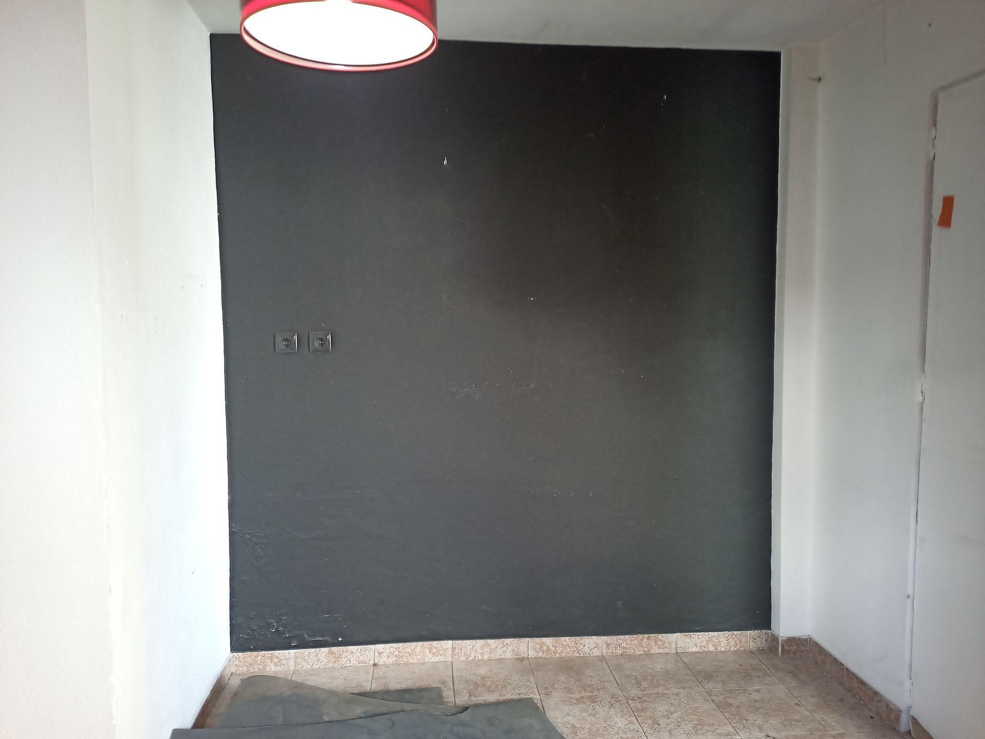 Parede pintada de preto com luminária, ladeada por paredes brancas e piso de azulejo.