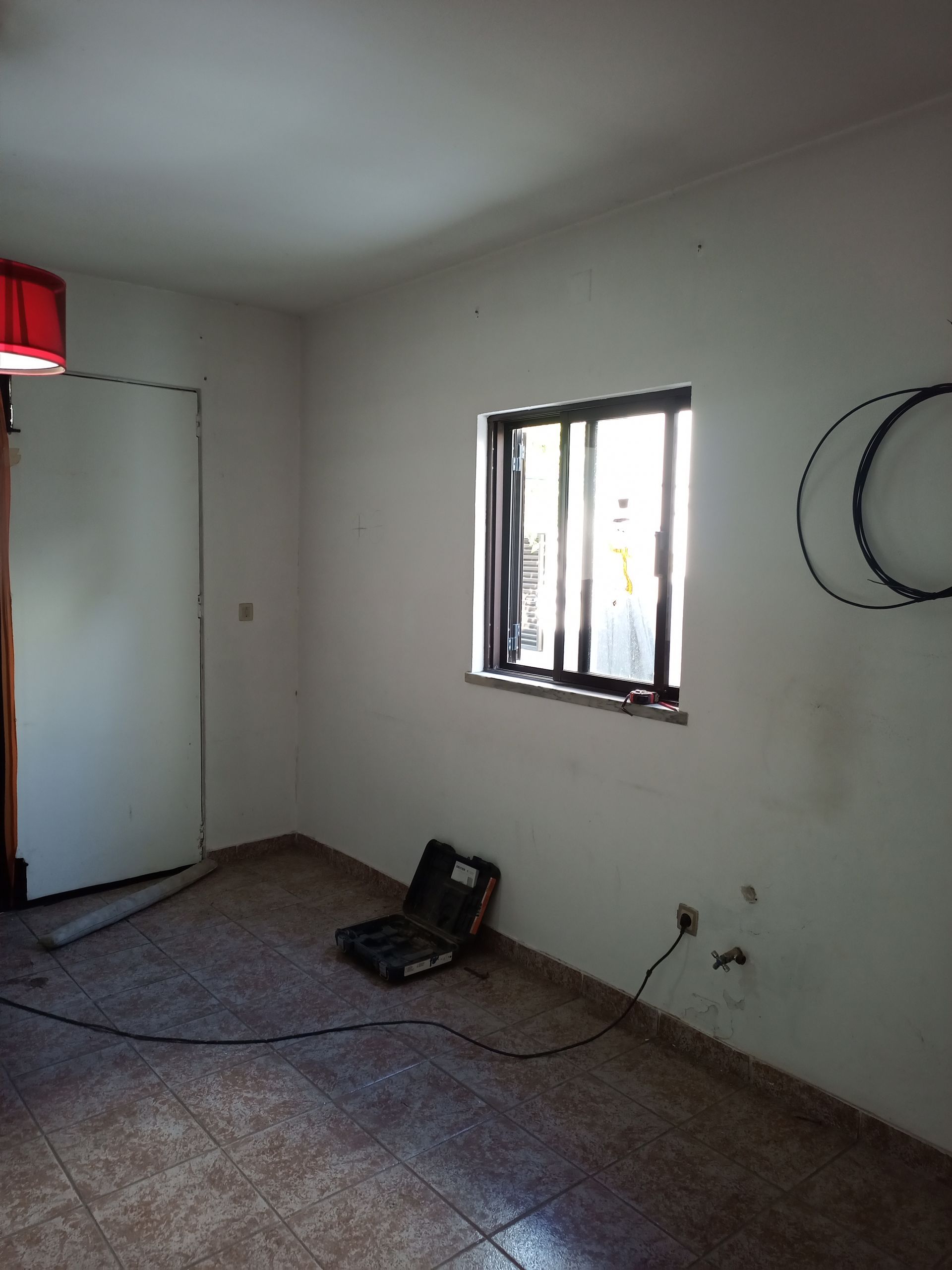 Quarto vazio com janela, porta e piso de azulejo; uma bolsa de ferramentas está perto da janela.
