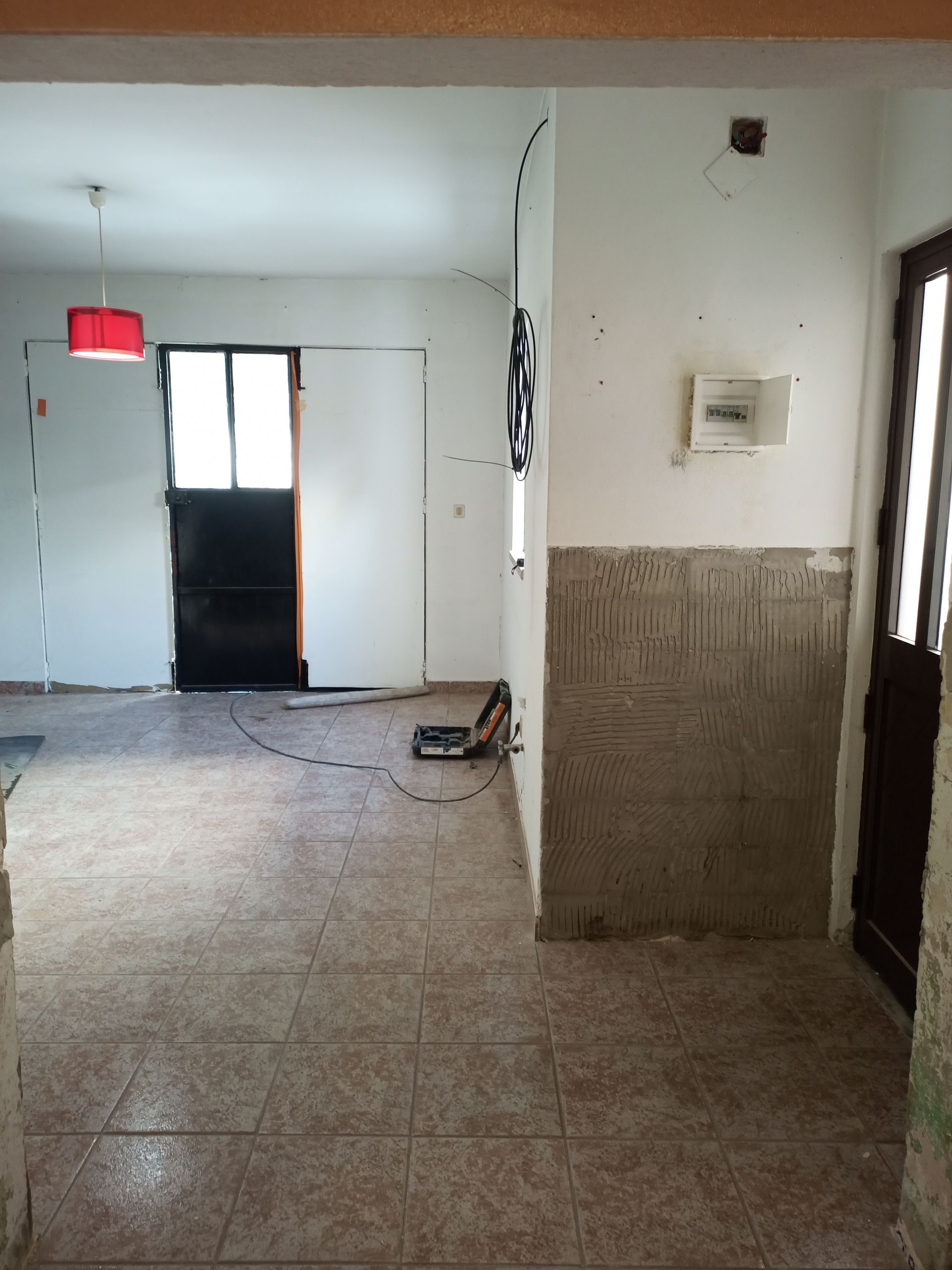 Vista interior de um quarto com piso de azulejo, porta aberta, luminária vermelha, porta preta e paredes brancas.