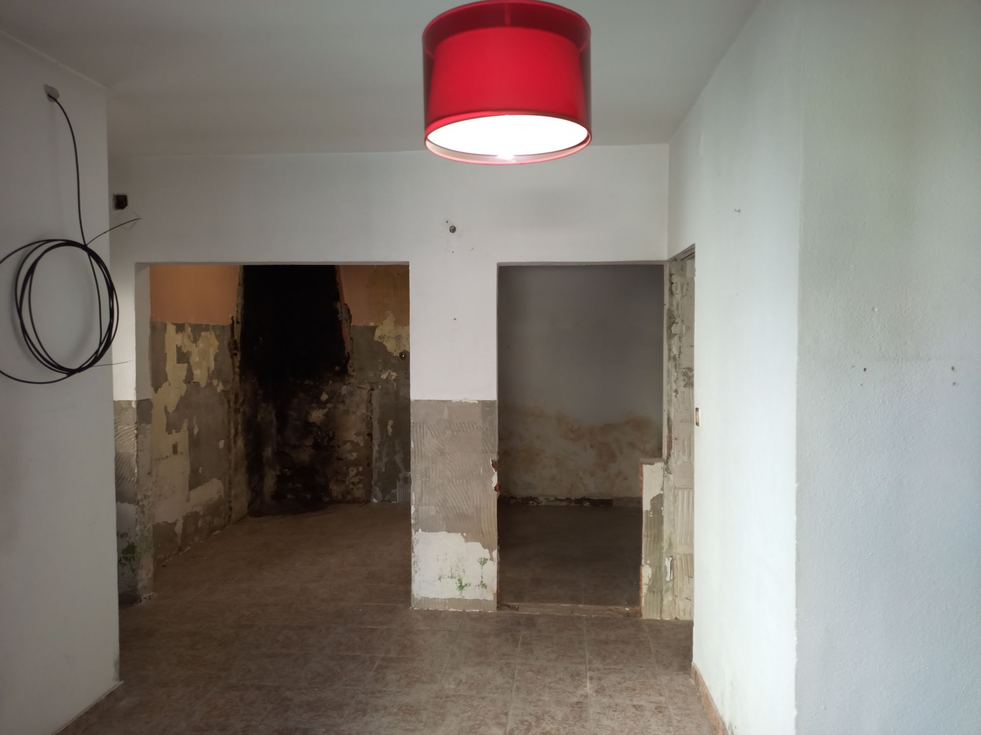 Vista interior de um cômodo com duas portas e uma luminária vermelha. As paredes apresentam tinta descascando e uma mancha escura.