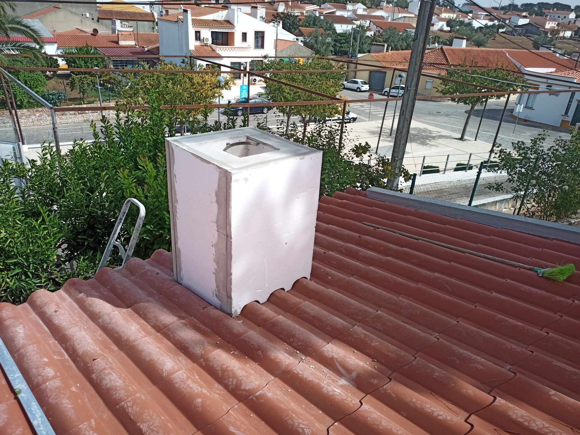 Chaminé branca sobre um telhado vermelho ondulado, rodeada por árvores e edifícios.