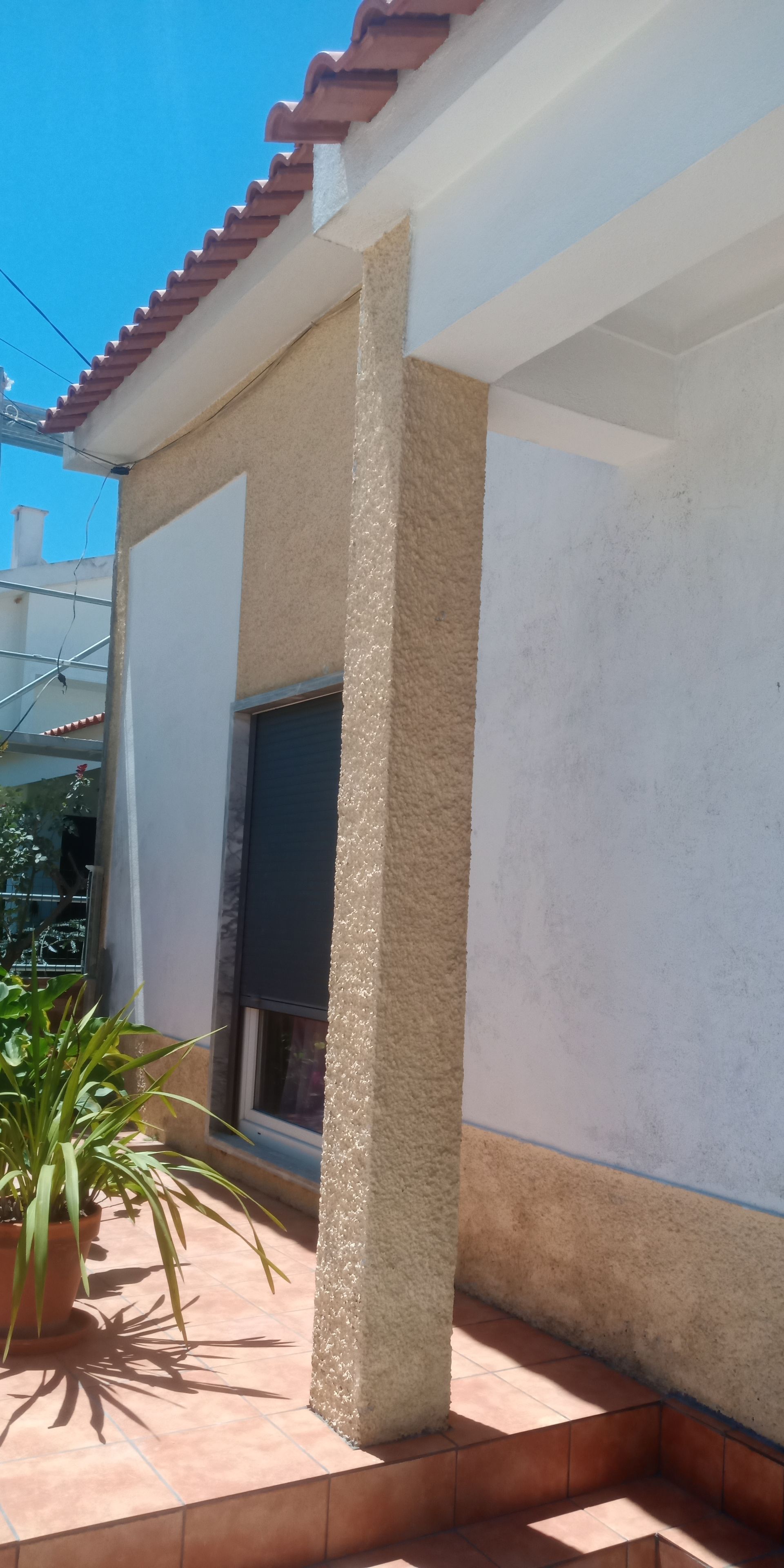 Vista exterior de um edifício com uma coluna texturizada, uma janela com persiana fechada e piso de ladrilho vermelho sob um céu azul.