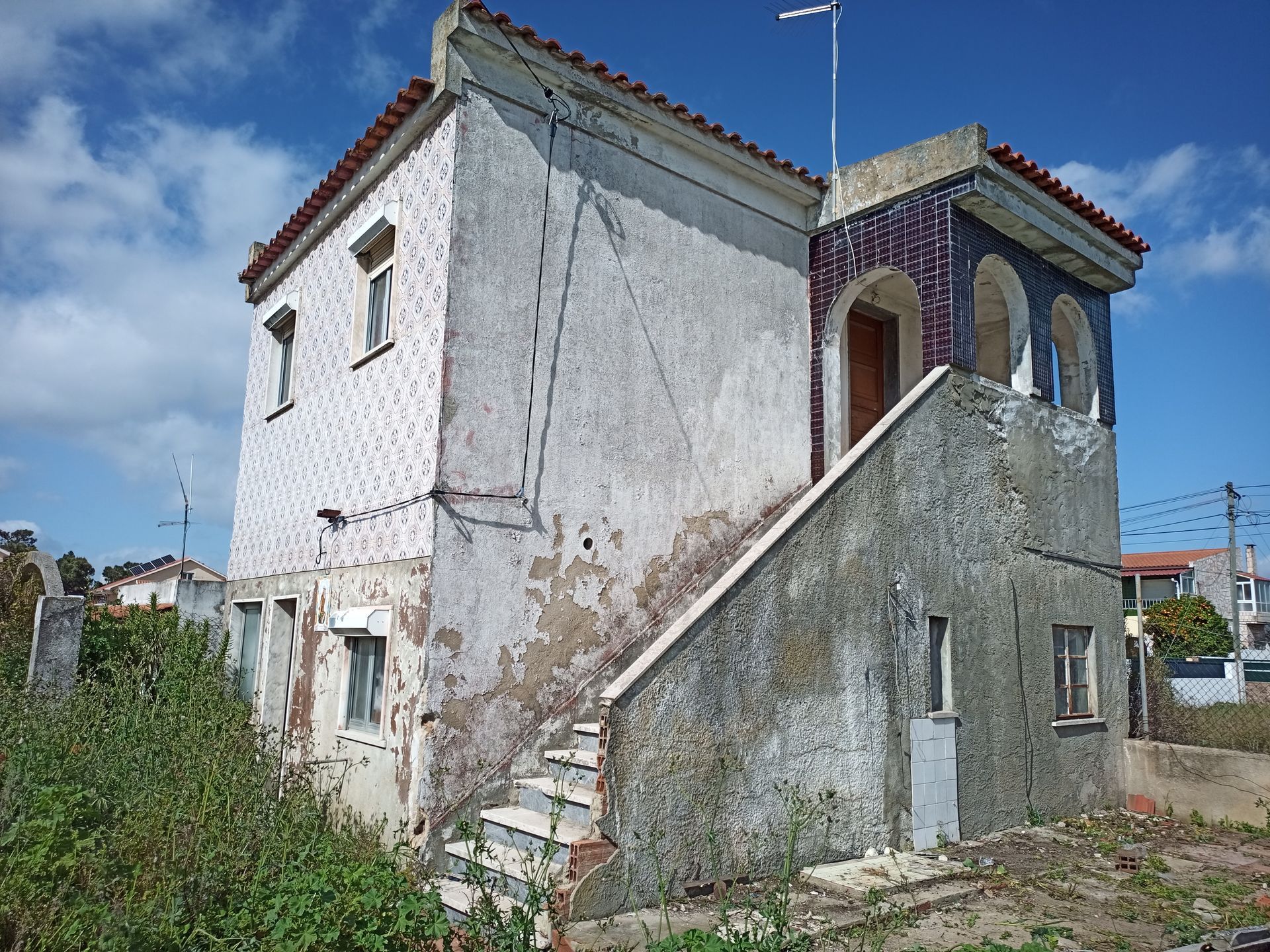 Prédio de dois andares com fachada desgastada e escada externa. Fachada branca com padrões, telhas vermelhas e céu azul ao fundo.