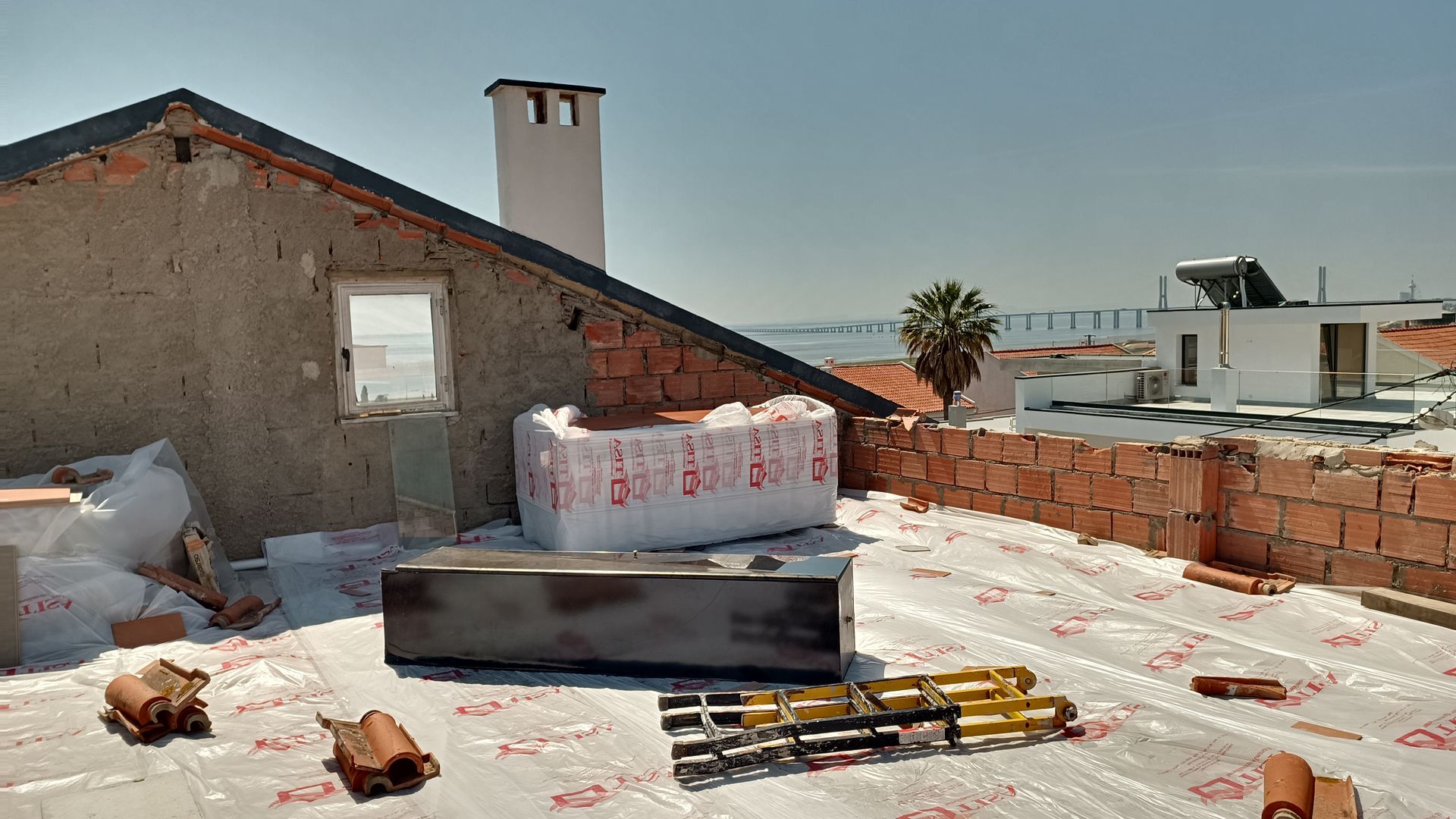 Construção no telhado: Isolamento, tijolos e um reservatório de água sobre uma camada de lona plástica. Céu azul ao fundo.