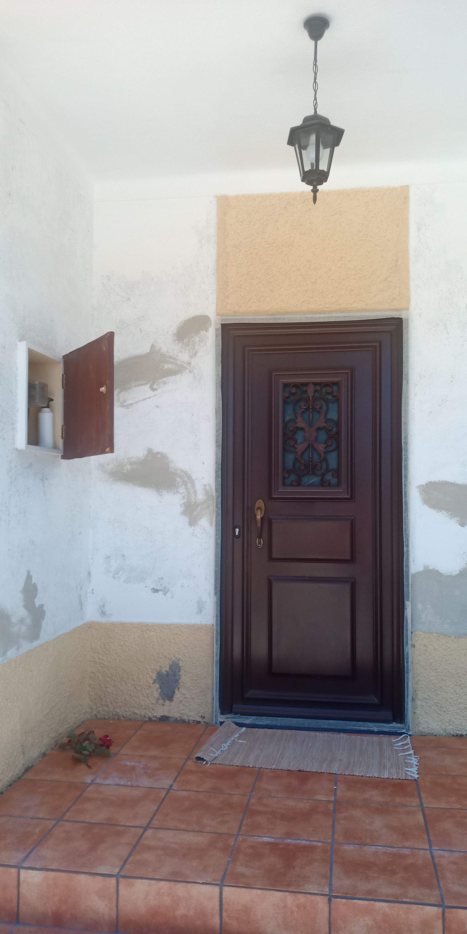 Uma porta de madeira marrom com um painel de vidro, sob uma luminária, em uma varanda com piso de azulejo.