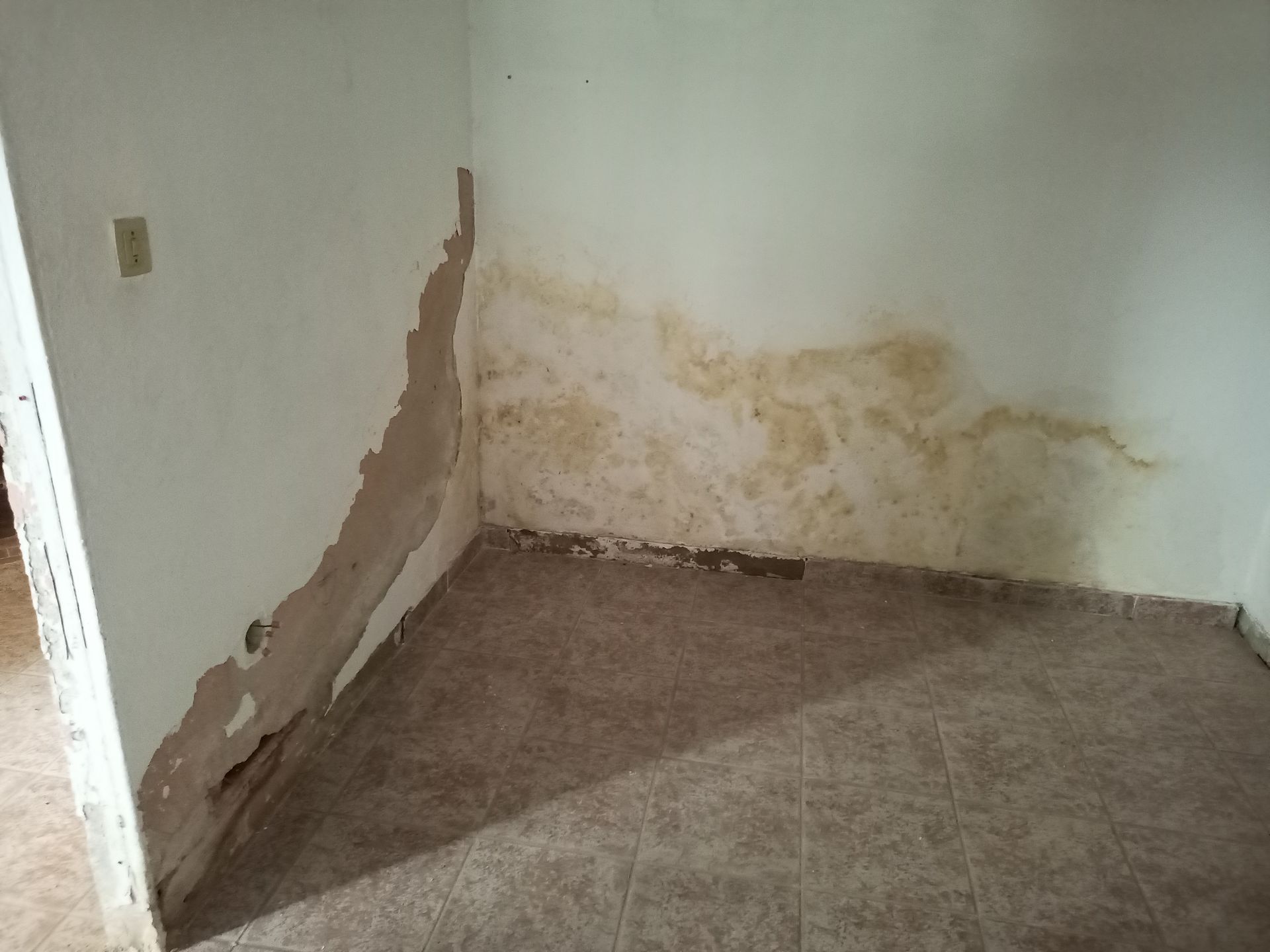Parede de canto interna com tinta descascando e danos visíveis causados ​​pela água, manchada com mofo marrom, sobre piso de azulejo.