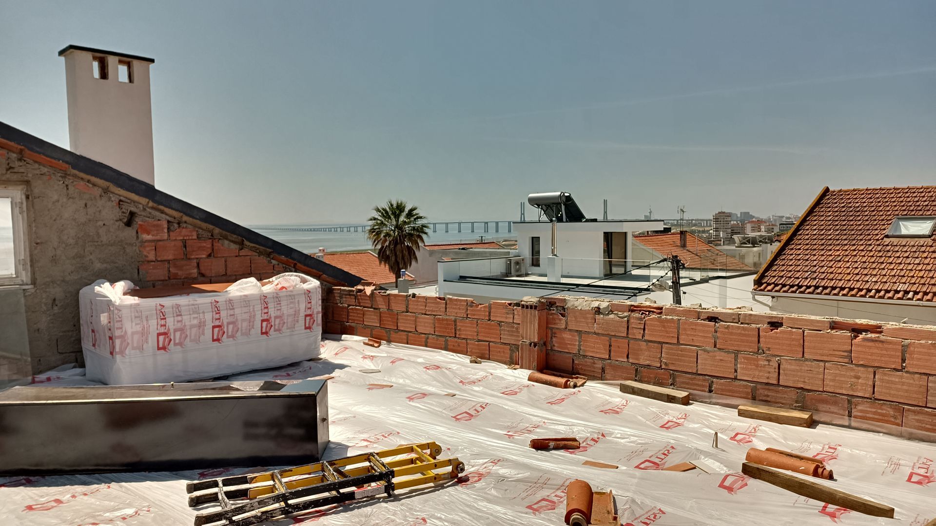 Telhado em construção com tijolos aparentes, lona plástica e uma ponte ao longe sob um céu claro.