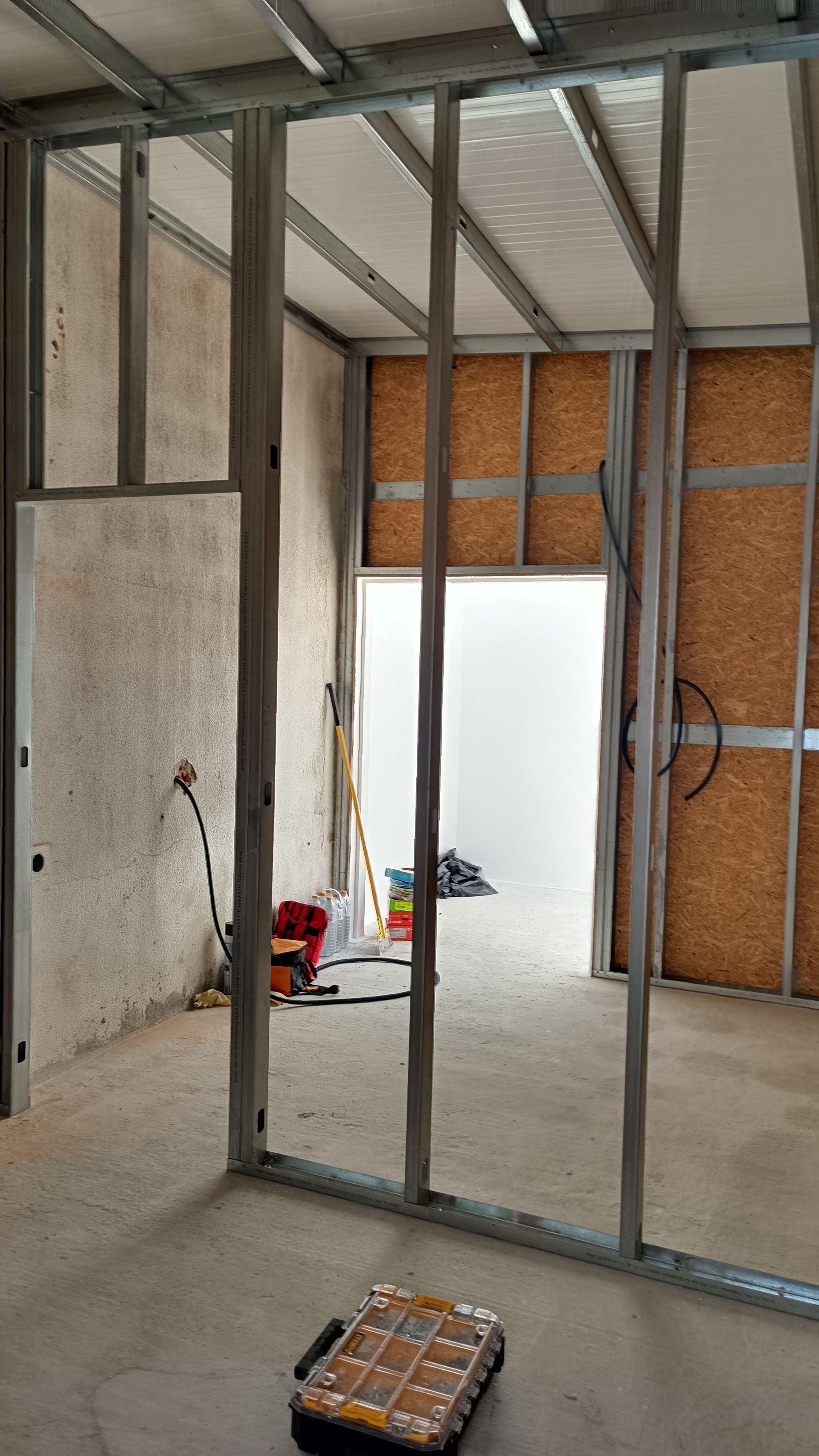 Construção interior: perfis metálicos estruturam as paredes com painéis de partículas e concreto aparente. Uma porta aberta permite a entrada de luz branca.