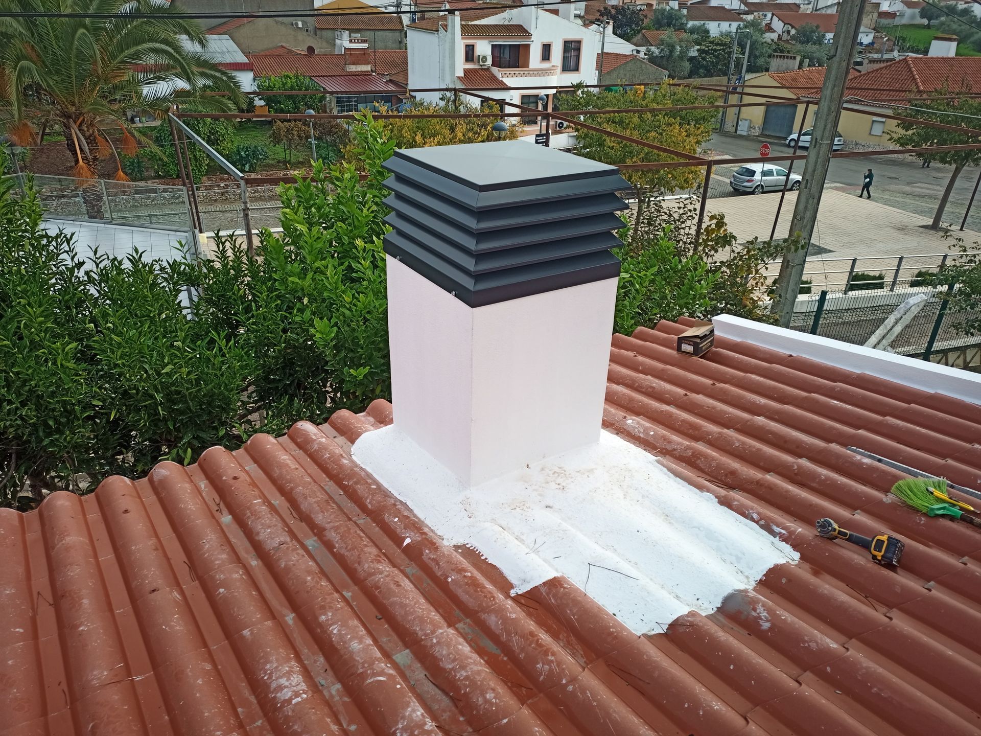 Chaminé em um telhado vermelho de zinco, cercada por vegetação e edifícios ao fundo.