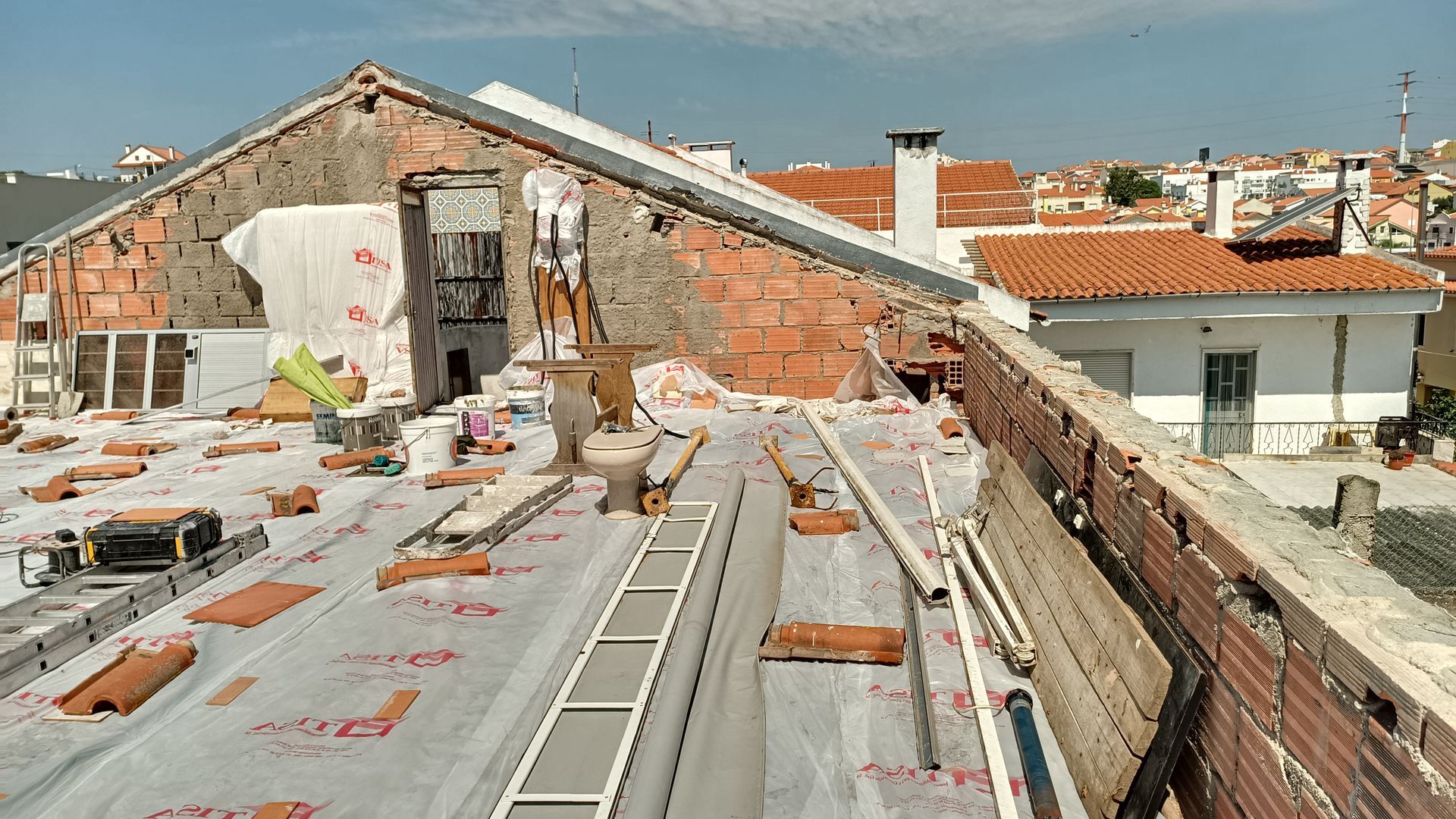 Telhado em construção. Tijolos aparentes, lonas e materiais de construção cobrem o telhado. Prédios ao fundo.
