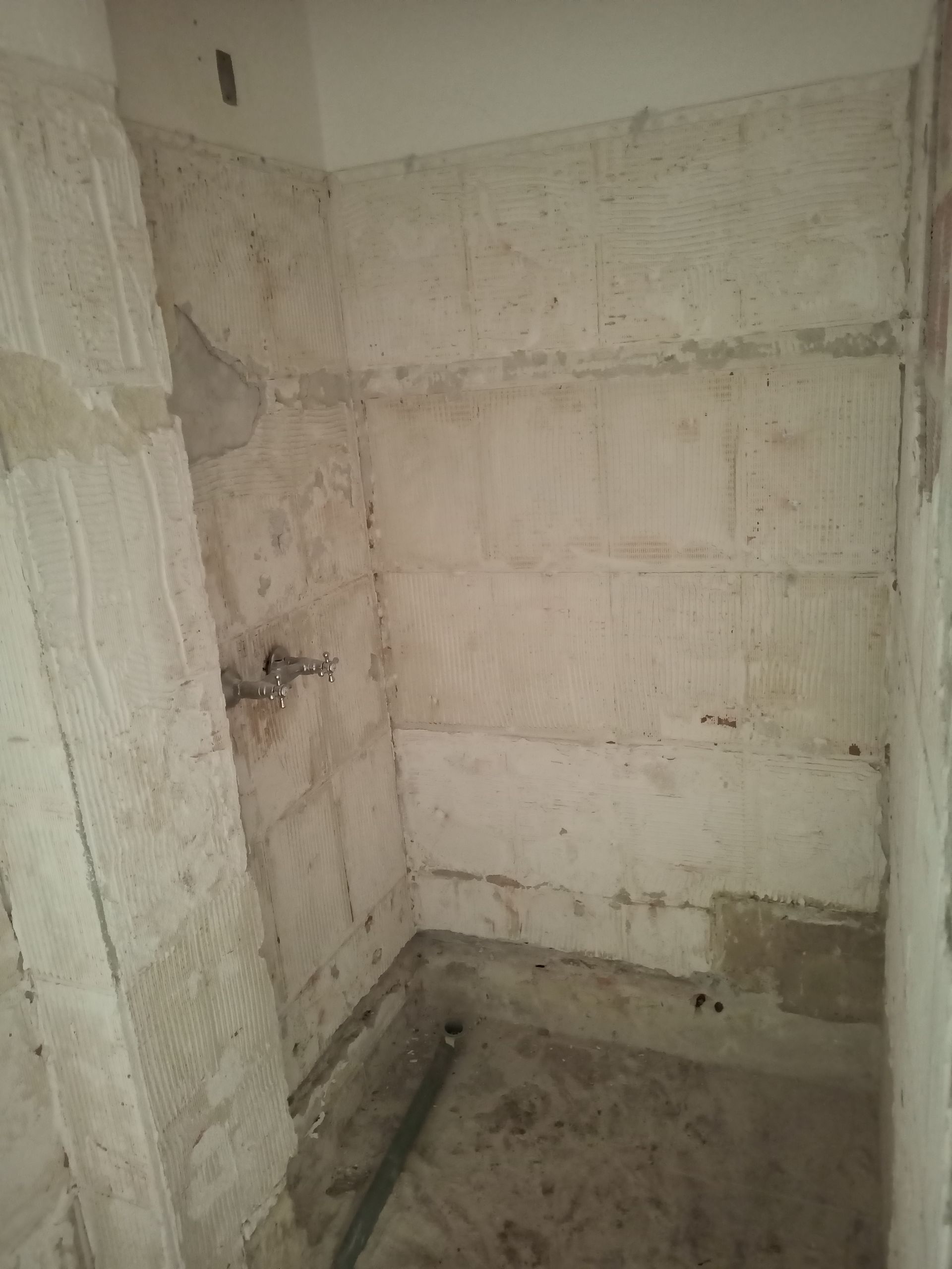 Banheiro com parede aparente, chuveiro exposto e piso de cimento. As paredes e o piso precisam de reparos/reformas.