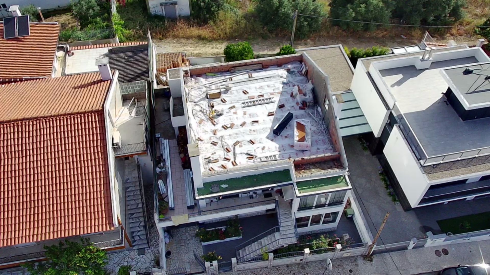 Vista aérea de um edifício em construção, com superfícies brancas, casas ao redor e um ambiente ensolarado.