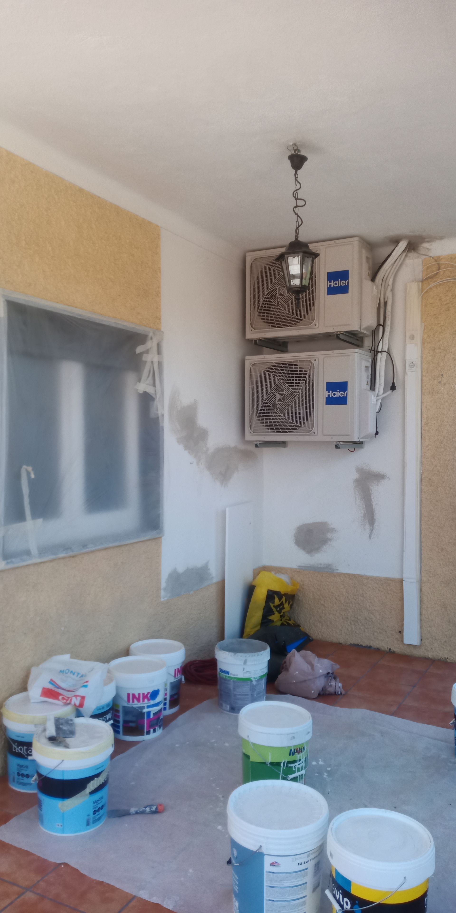 Vista interna de uma varanda com baldes de tinta, aparelhos de ar condicionado e uma janela coberta com lona plástica.