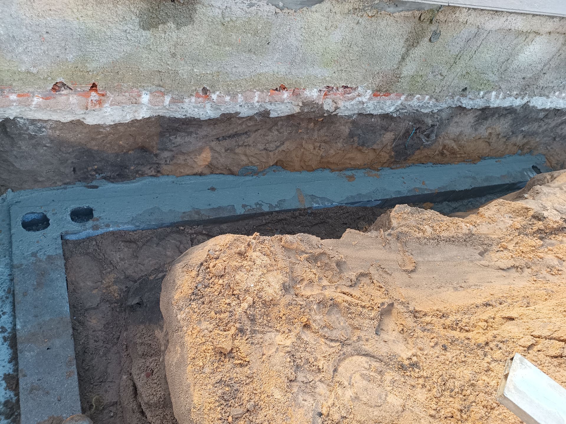 Valeta escavada ao longo de uma fundação de concreto. Parte do concreto foi despejada no fundo, com solo e tijolos expostos.