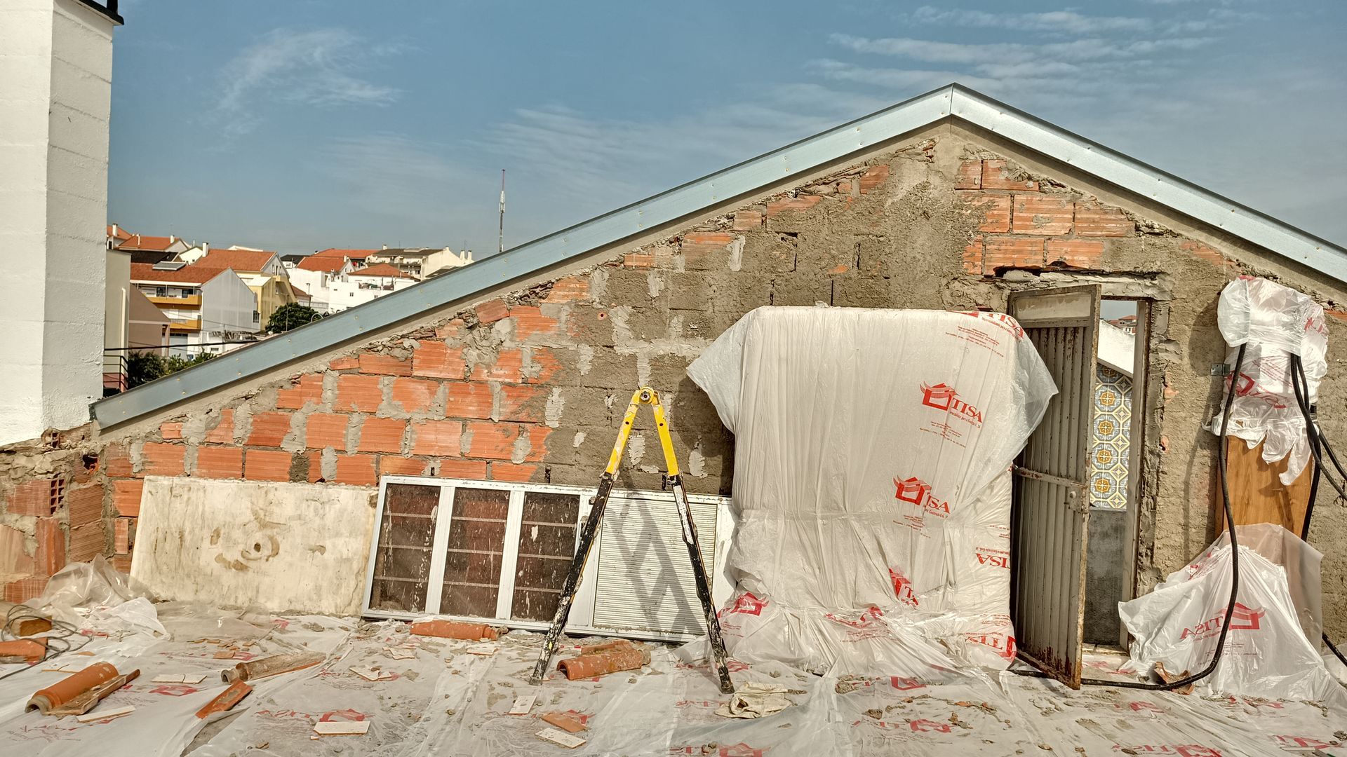 Telhado em construção, parede de tijolos aparentes, acabamento metálico no telhado, lona plástica, escada, luz solar.