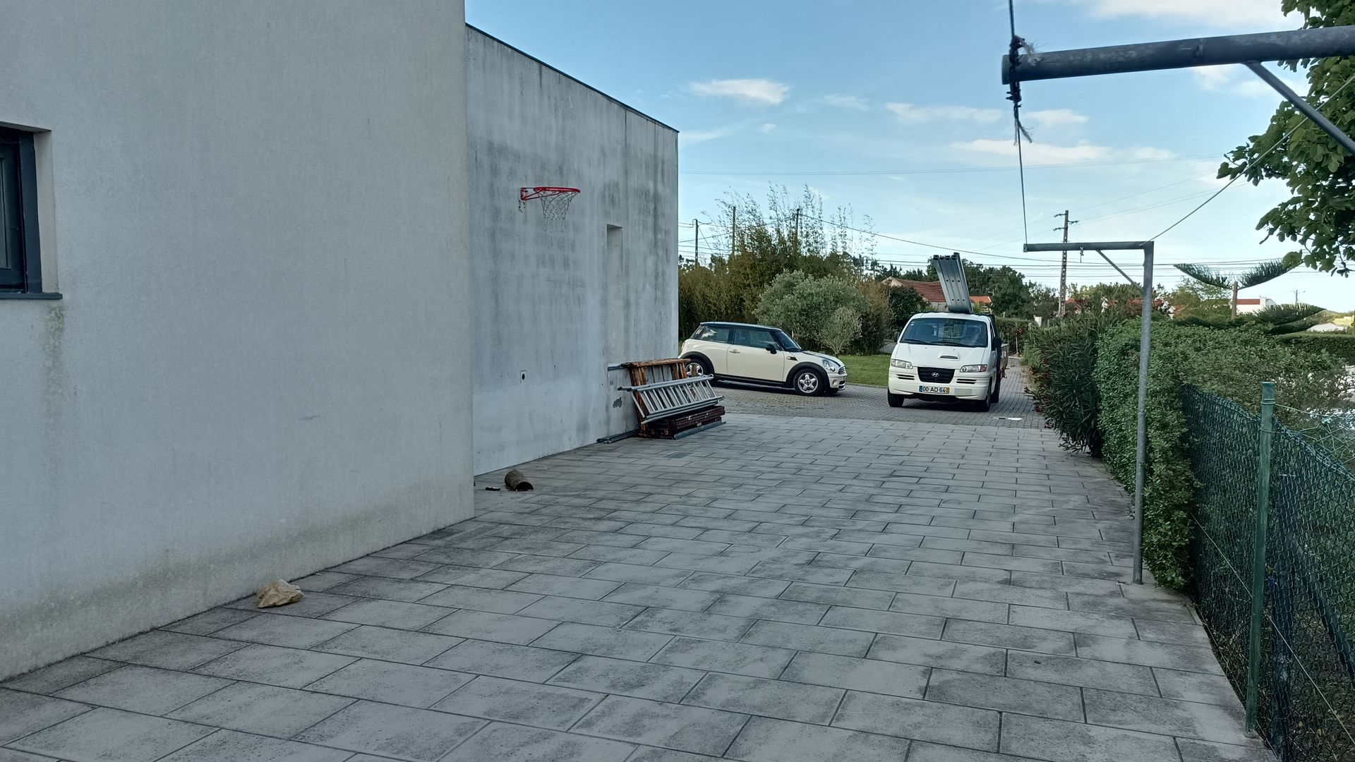 Prédio branco com uma cesta de basquete, carros estacionados em uma entrada pavimentada, cerca verde à direita.