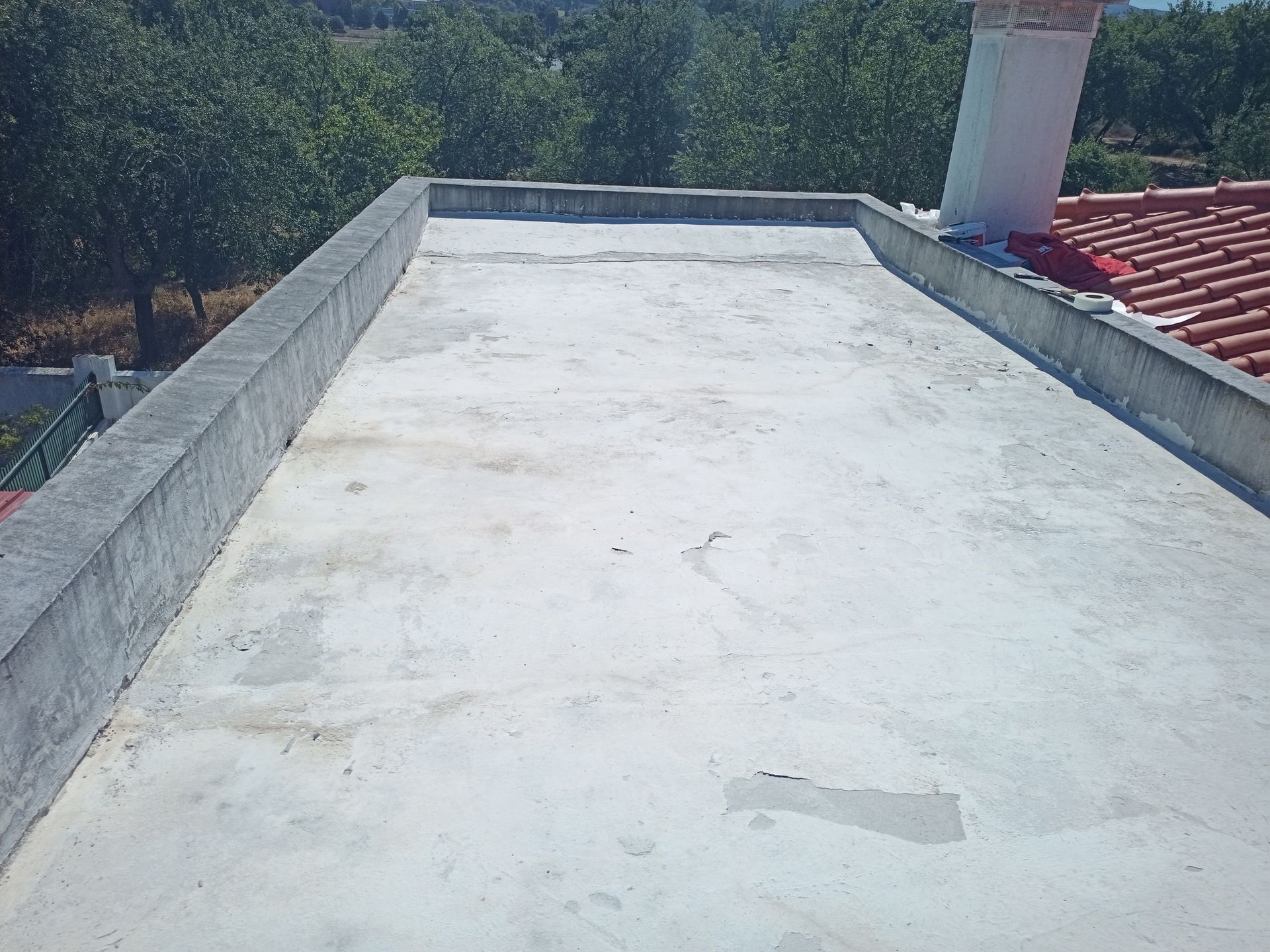 Telhado plano de concreto com muros baixos, chaminé e árvores ao fundo.
