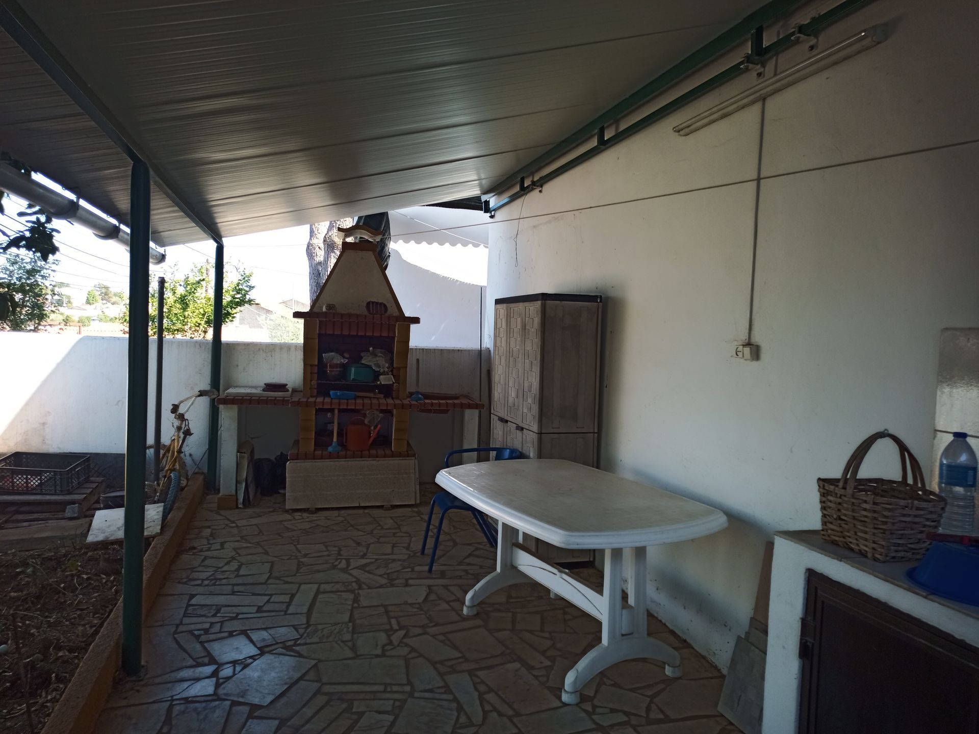 Varanda coberta com churrasqueira, mesa e armário para armazenamento. Paredes claras e piso de azulejo.