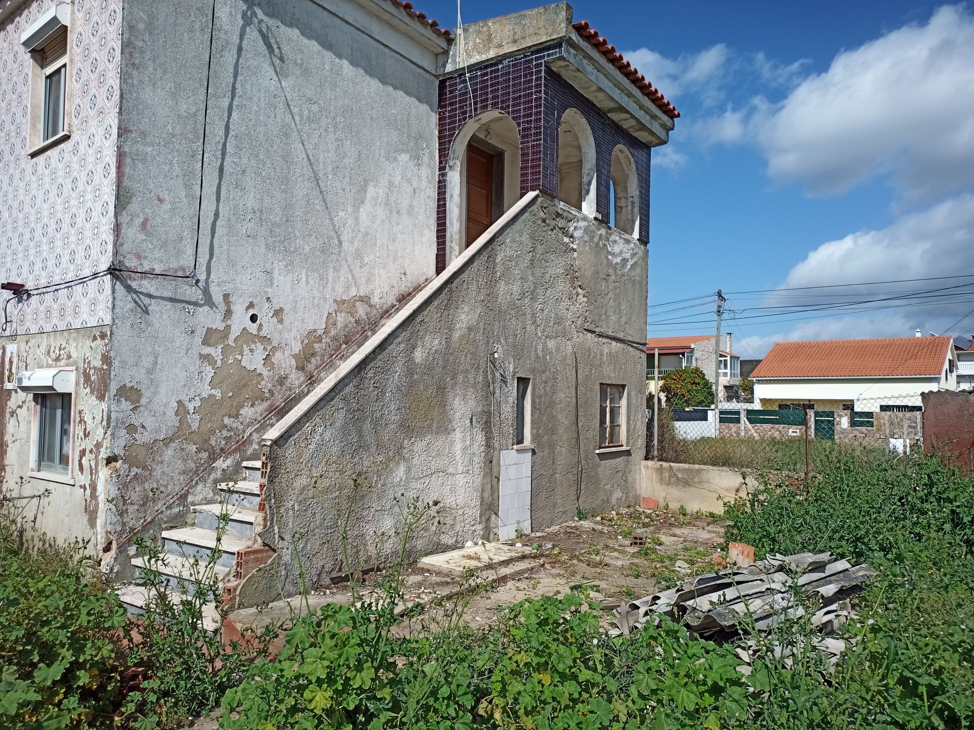 Exterior de um prédio de dois andares desgastado pelo tempo, com uma escadaria e uma pequena varanda revestida de azulejos. Vegetação densa o cerca.