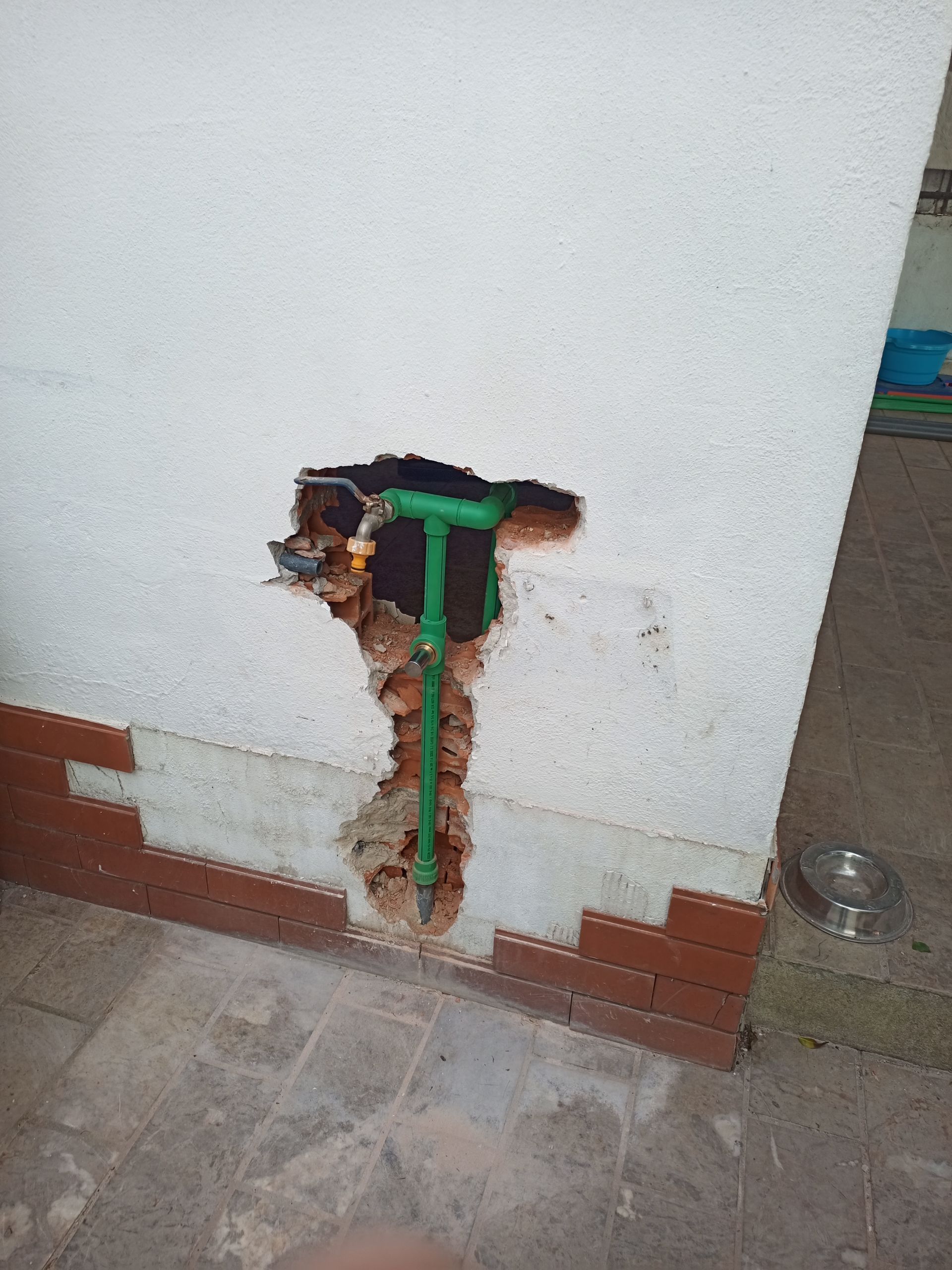 Buraco em uma parede branca expondo encanamento verde e alvenaria. A parede encontra uma borda e um piso que imitam tijolos.