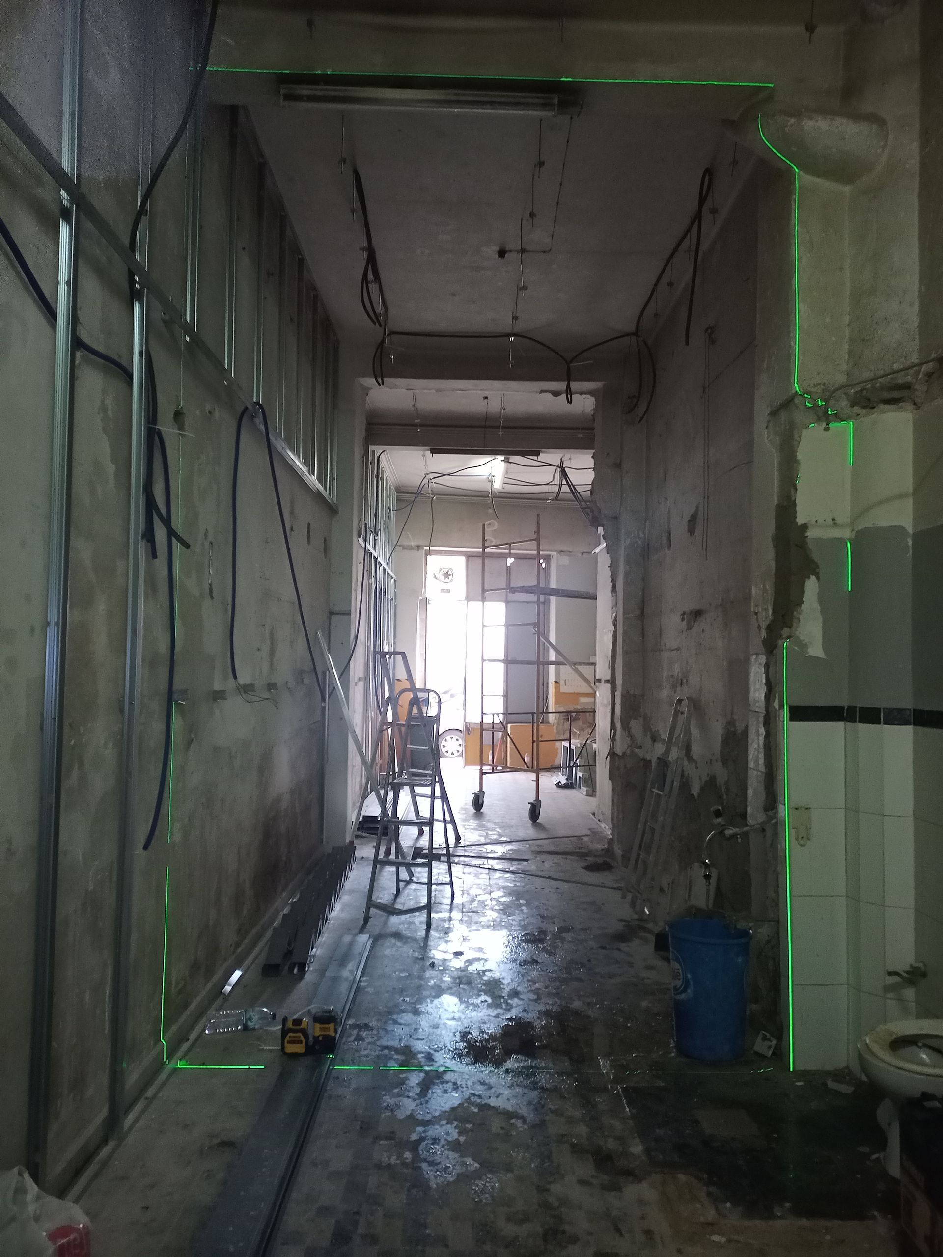 Corredor reformado com linhas verdes de laser, vigas metálicas, fiação exposta e equipamentos de construção.