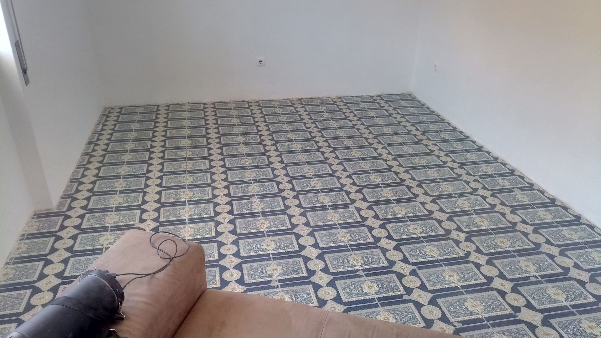 Quarto com carpete estampado em tons de azul e bege, paredes brancas. Sofá bege em primeiro plano.