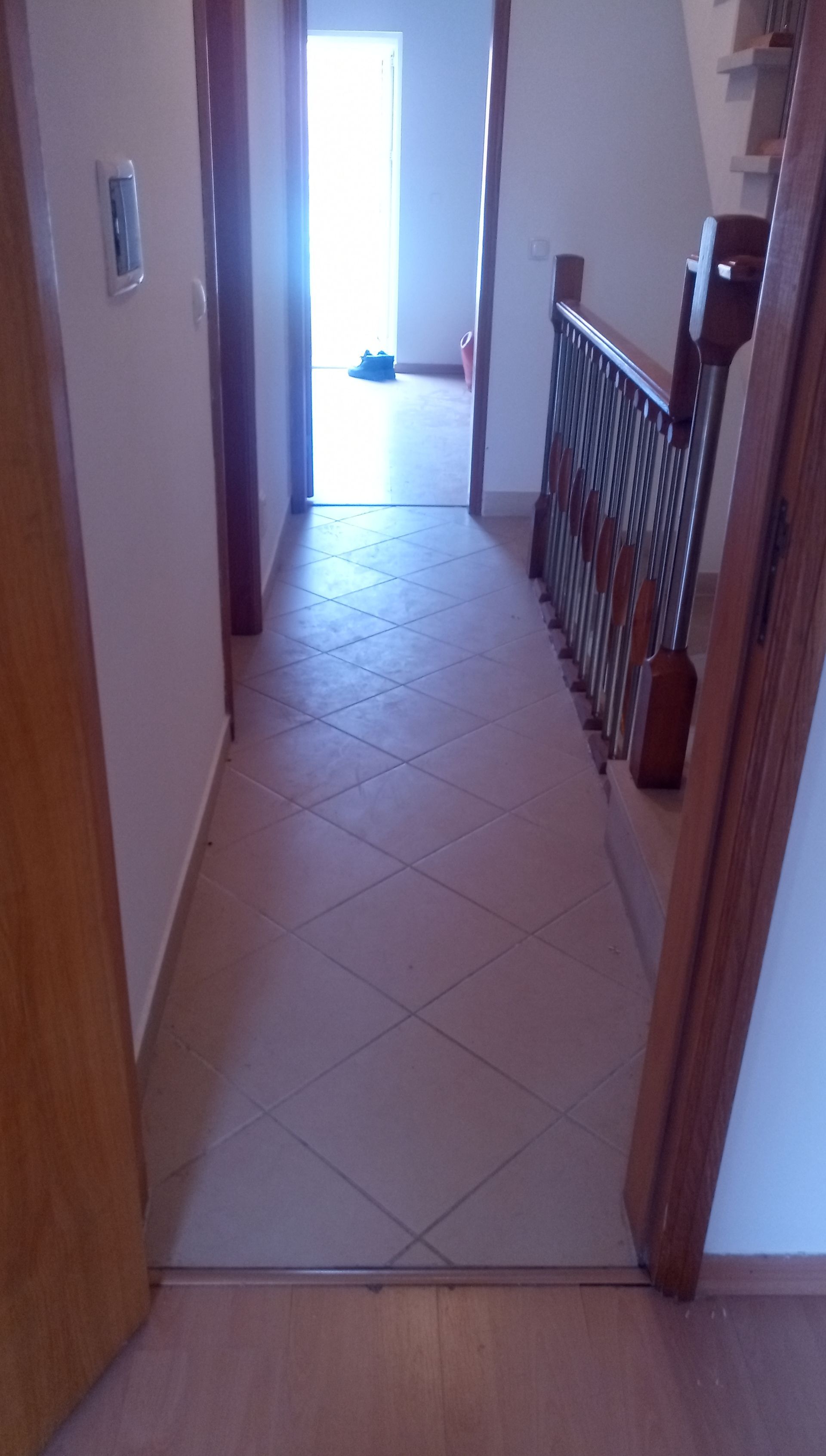 Corredor com piso de azulejo, portas abertas e uma escada com corrimão decorativo. Iluminação abundante no final.