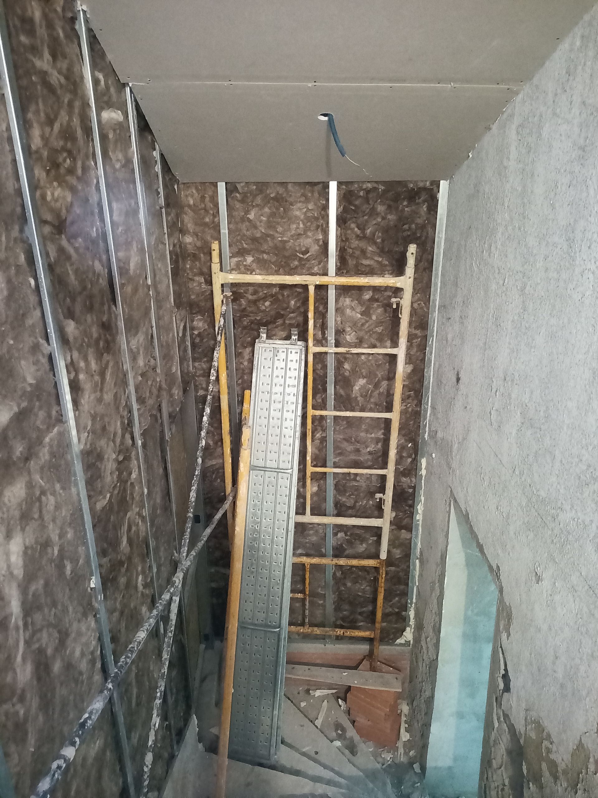 Escadaria em construção; isolamento exposto, andaimes, teto de gesso acartonado, paredes de concreto.