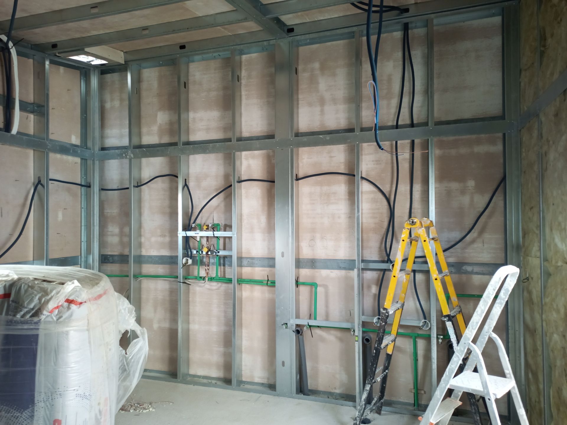 Quarto interior em construção, paredes com estrutura metálica, instalações hidráulicas e elétricas visíveis, escada no canto.