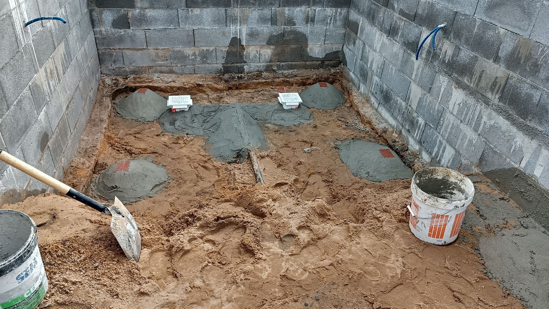 Canteiro de obras. Uma sala retangular com paredes de blocos de concreto. Cimento está sendo despejado ao redor de canos de drenagem no chão.