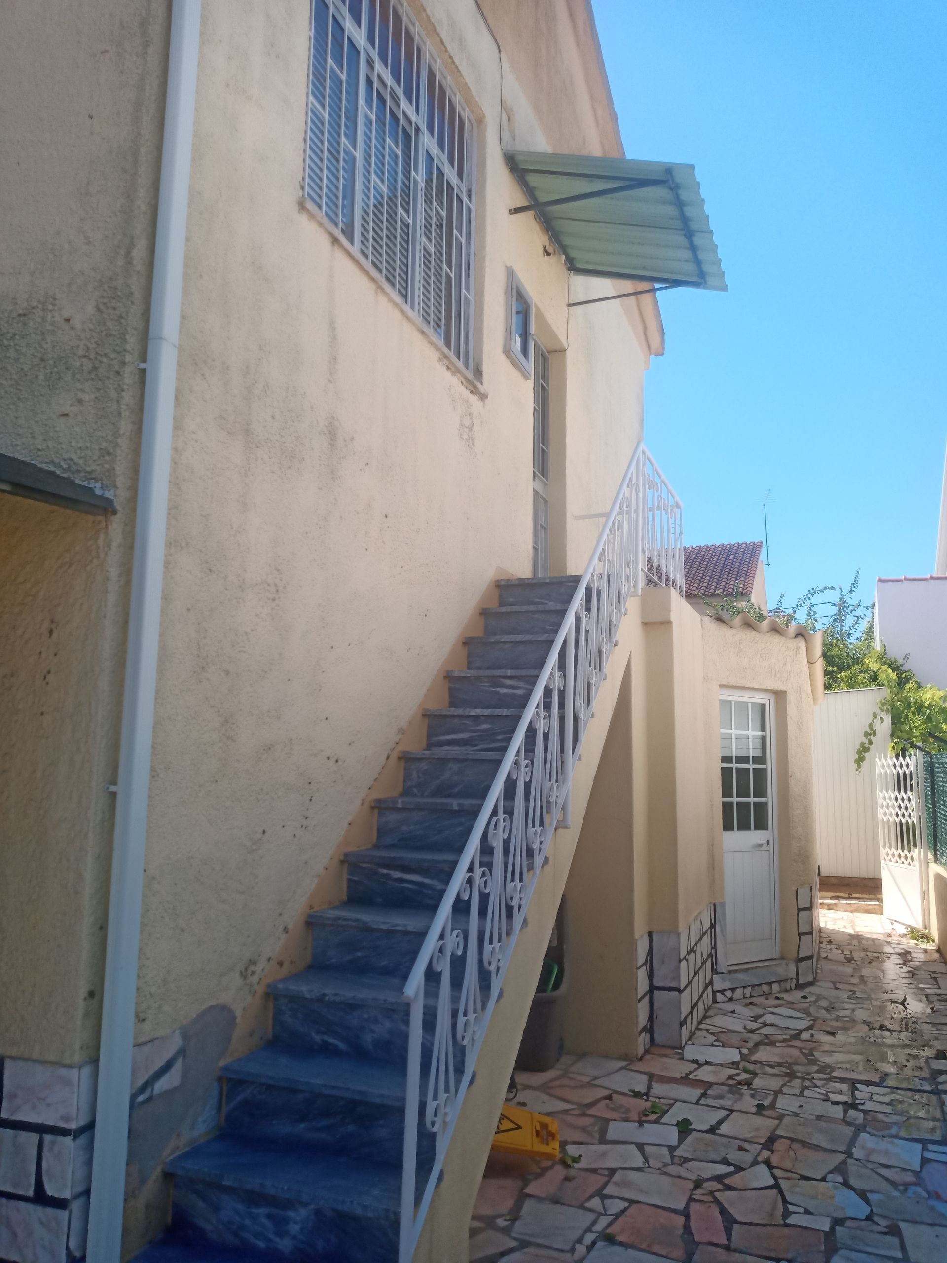Vista exterior de um edifício com escadaria de pedra que conduz a uma porta e uma janela. Paredes de estuque amarelo claro e céu azul.