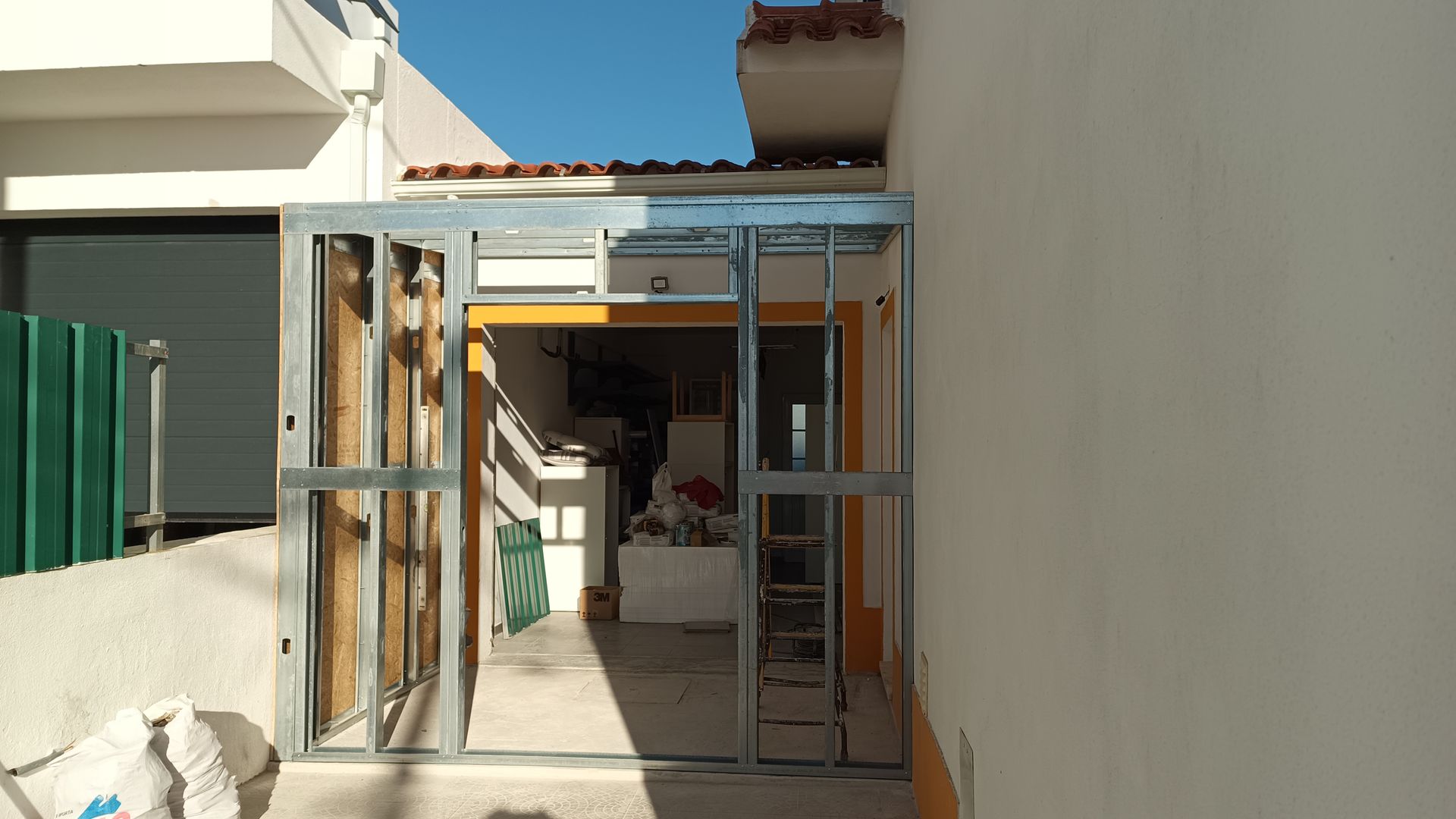 Porta com estrutura metálica em beco estreito. A luz do sol ilumina o interior. Paredes brancas do prédio.