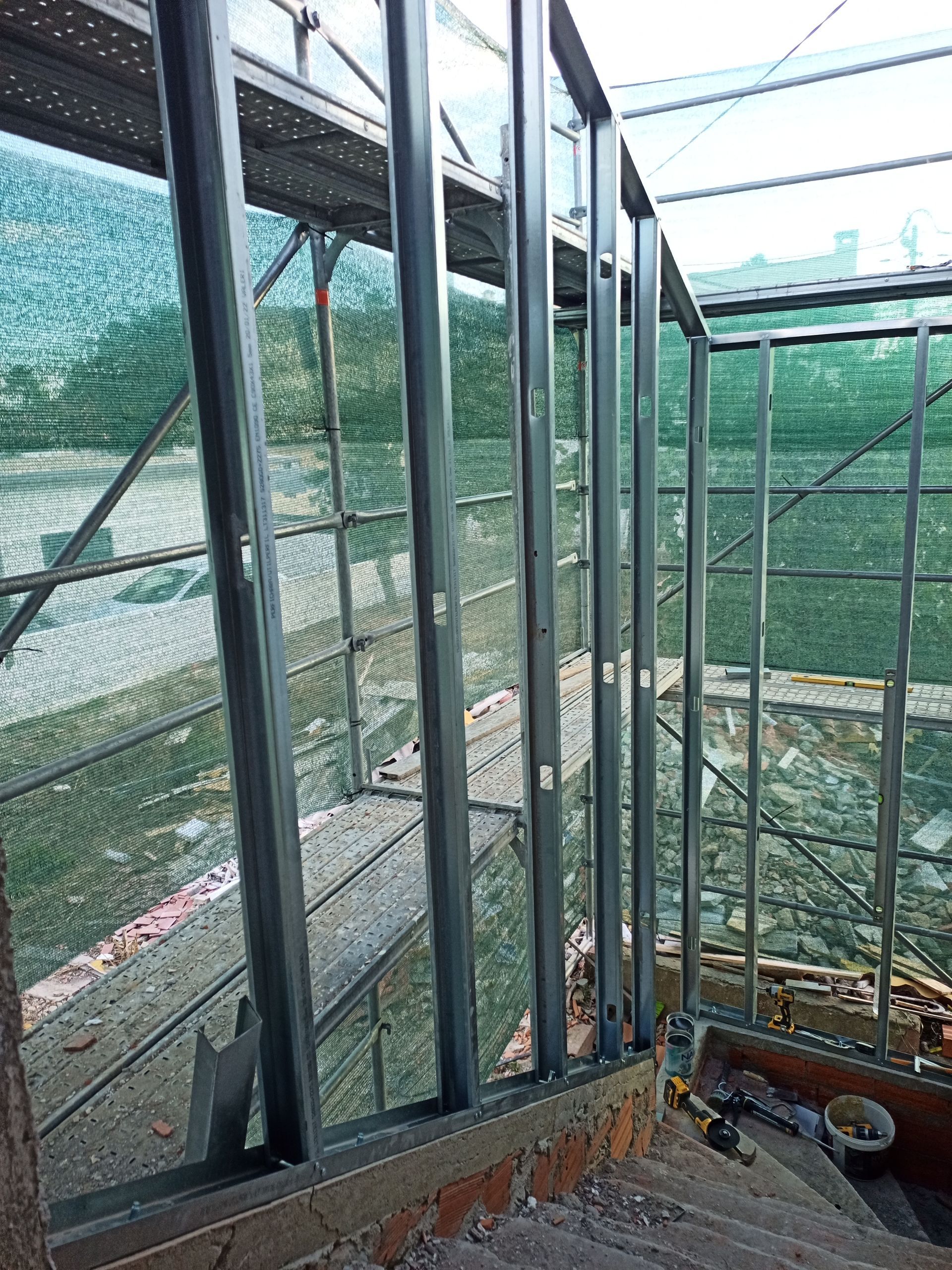 Estrutura de aço que forma uma parede, possivelmente uma escada, em área externa. Rede verde ao fundo.