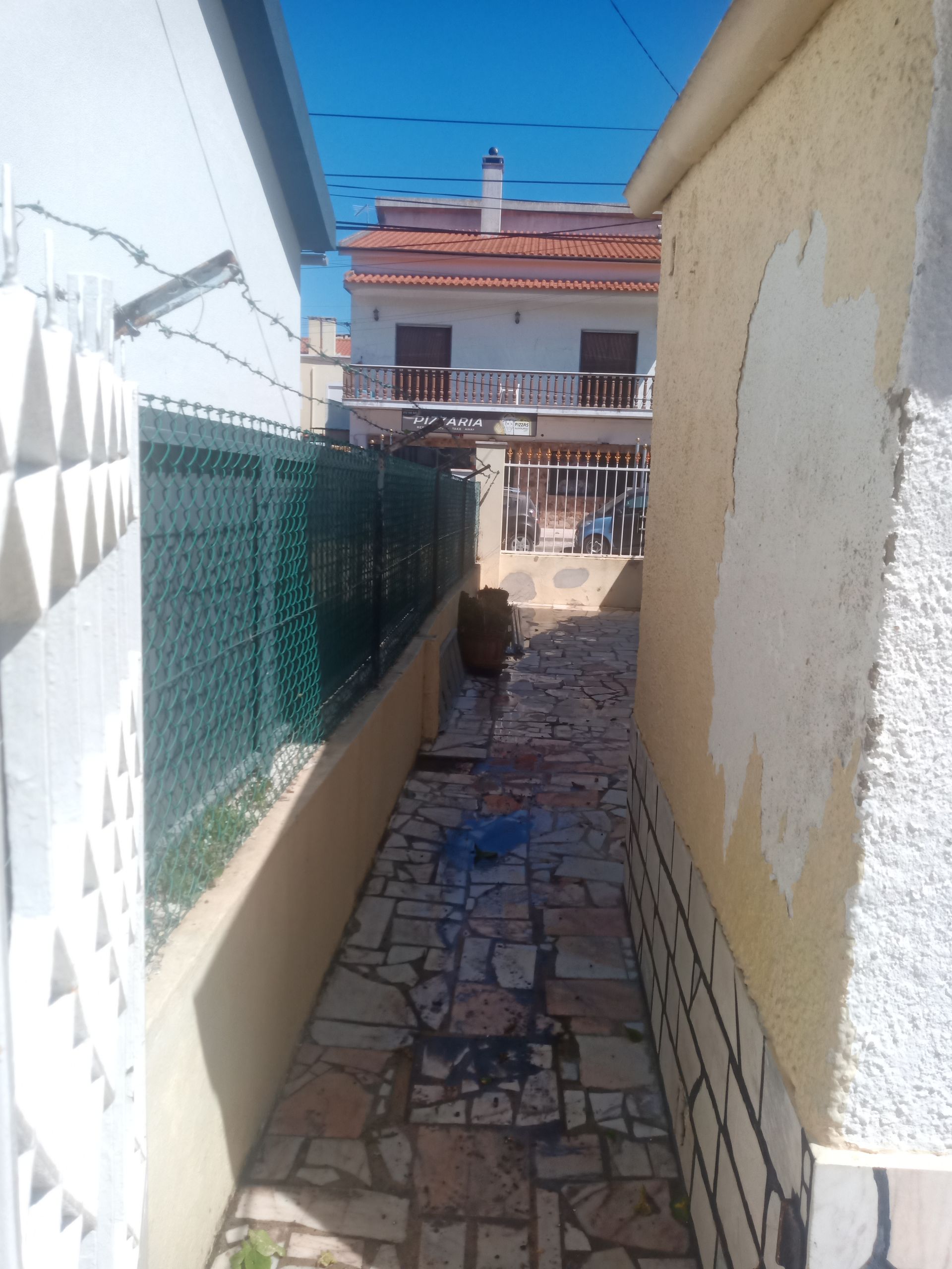 Caminho estreito de pedra entre dois edifícios; cerca verde de um lado, edifício com varandas ao longe.
