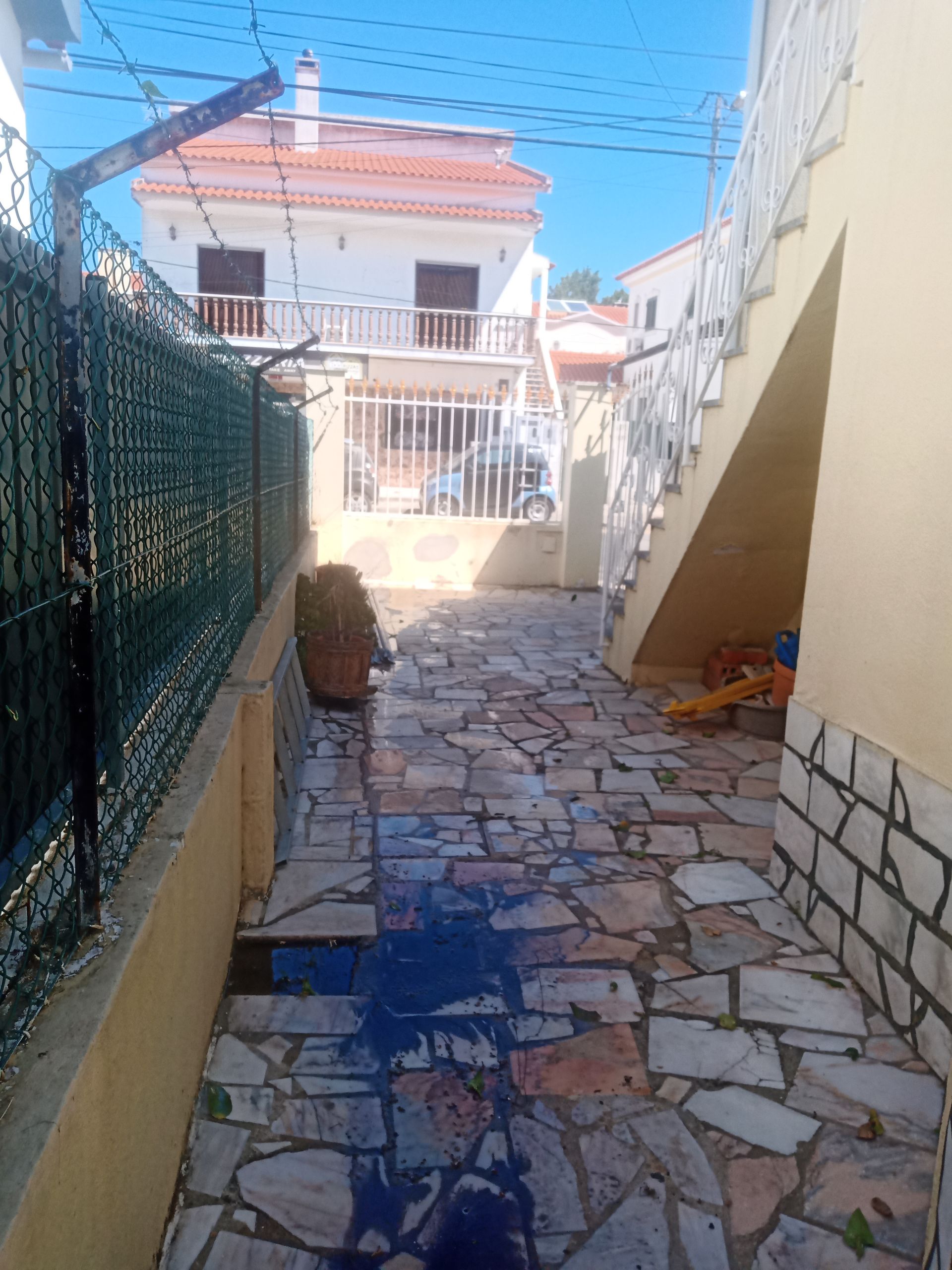 Uma estreita passarela de pedra entre uma cerca verde e uma escadaria, que leva a edifícios sob um céu claro.