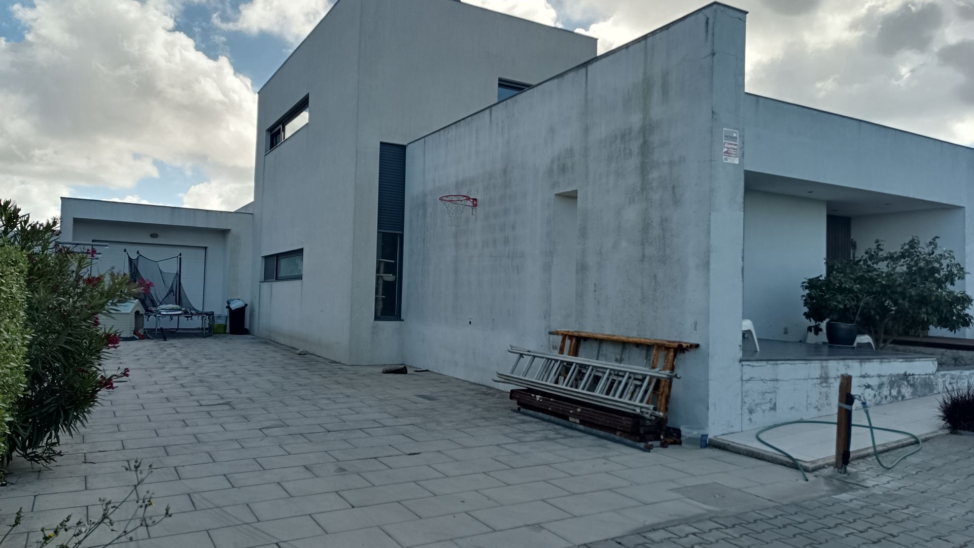Prédio moderno branco de dois andares com entrada de concreto e céu nublado. Escada e cesta de basquete visíveis.