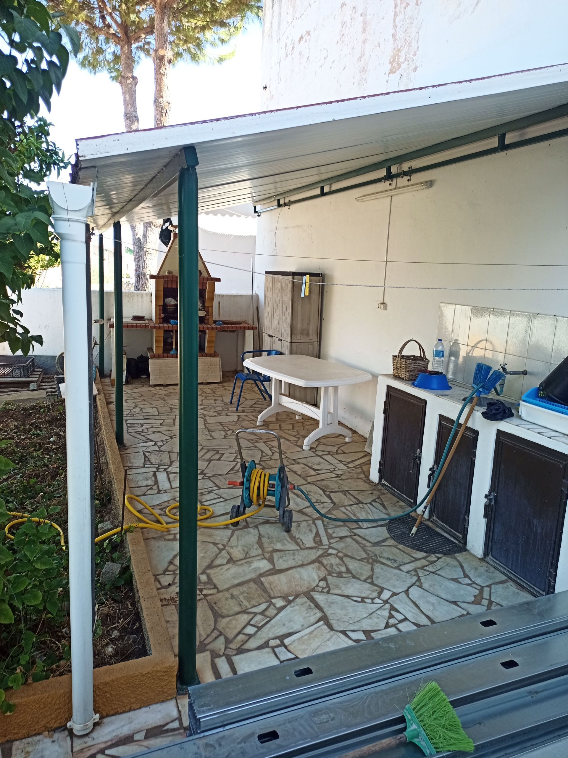 Cozinha externa coberta com armários brancos, mesa e churrasqueira. Pilares verdes sustentam um telhado branco.