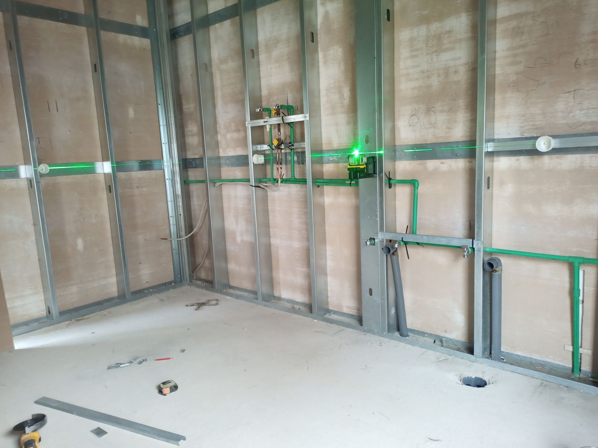 Banheiro em construção: estrutura metálica, tubulações verdes, nível a laser e paredes inacabadas.