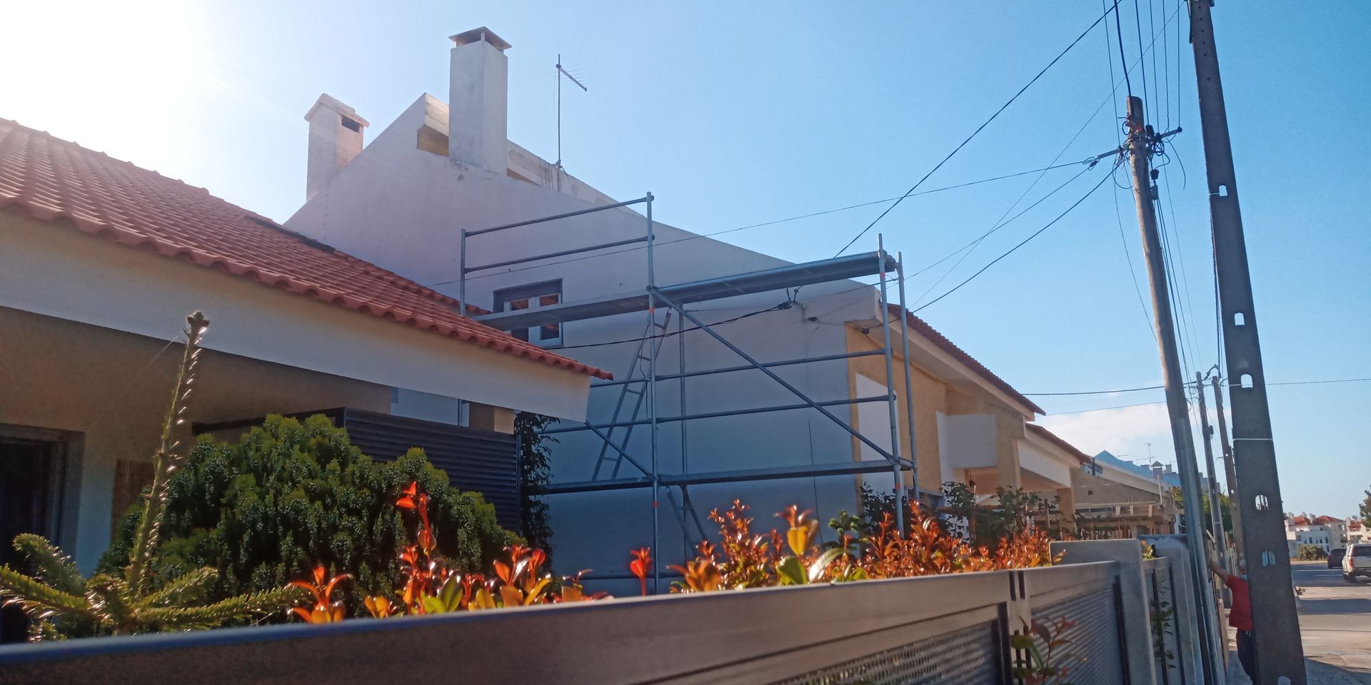 Exterior de uma casa com andaimes, telhado de telha vermelha e fios de energia elétrica contra um céu azul claro.