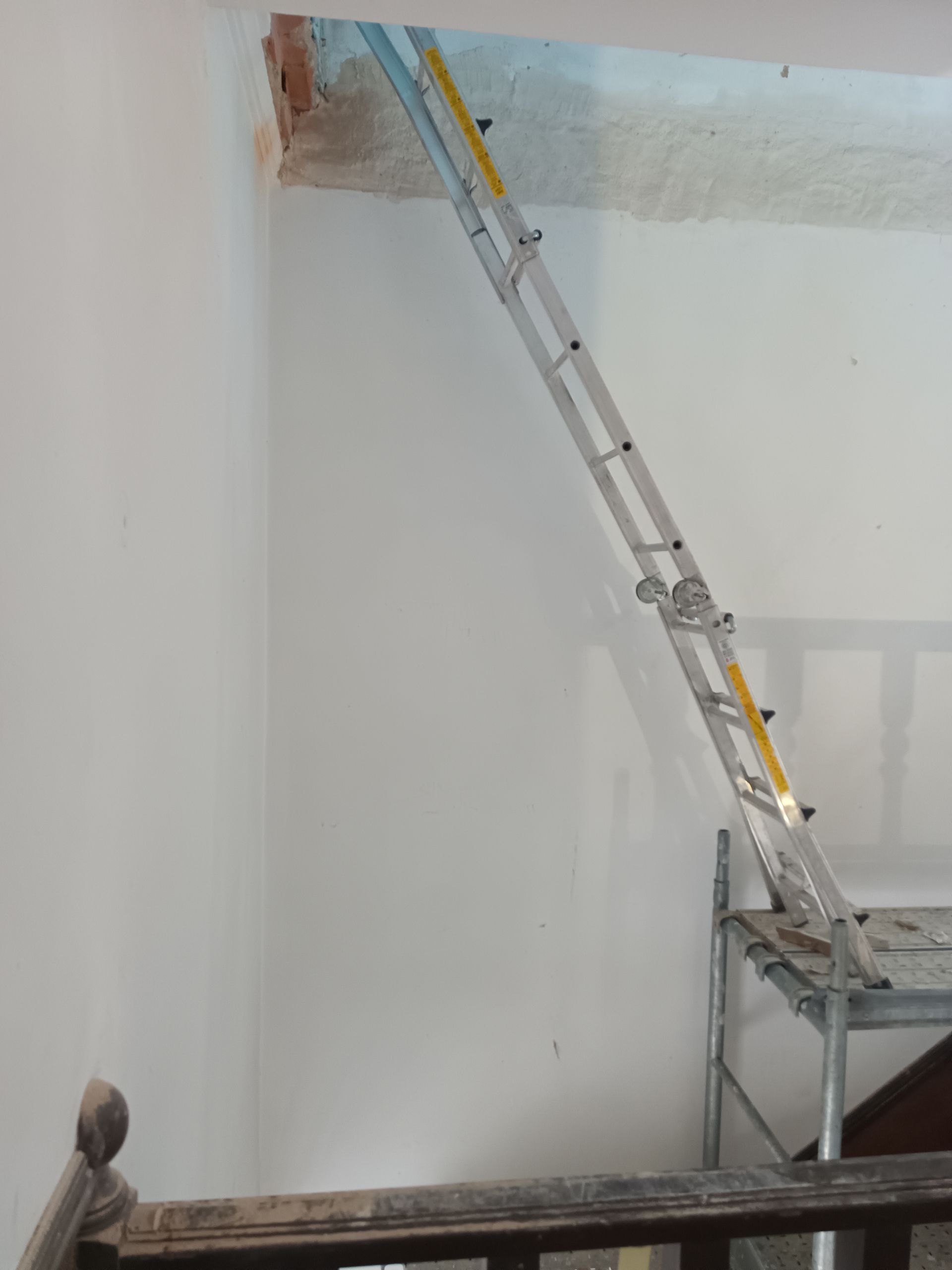 Escada encostada na parede, alcançando um teto danificado. Andaimes são visíveis na base.