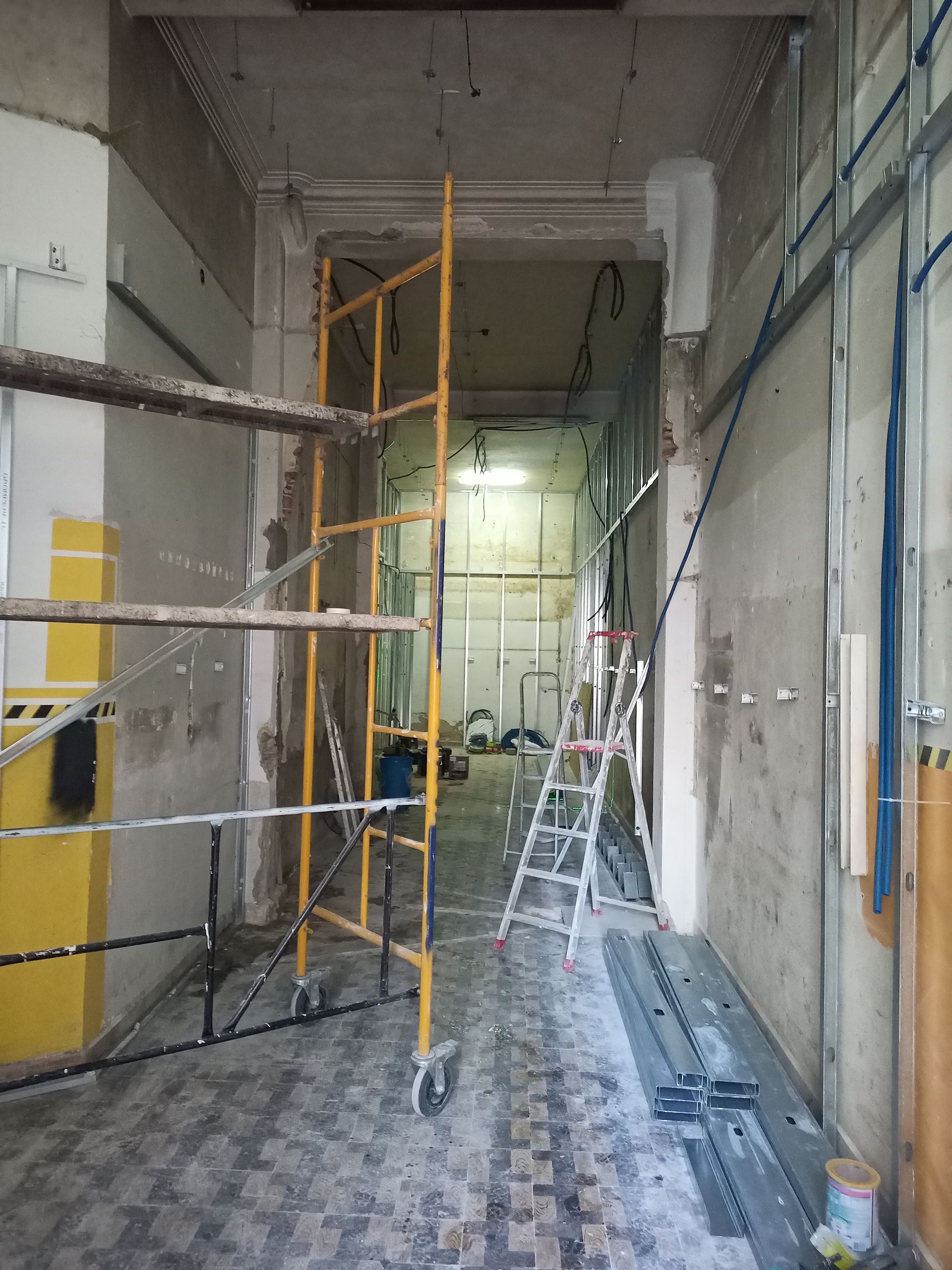 Corredor em construção com andaimes e escadas. Paredes cinzentas, teto ornamentado, estrutura metálica azul.