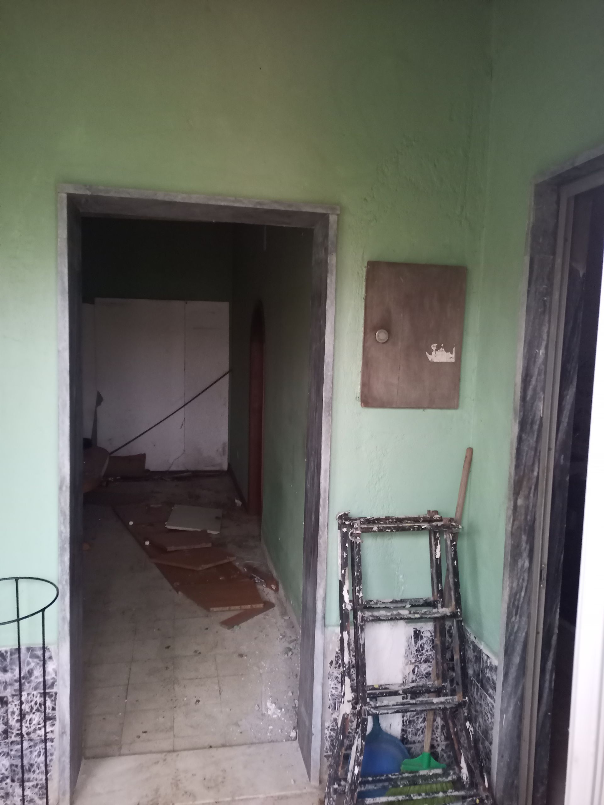 Uma porta com paredes verdes dá acesso a um corredor desarrumado. Detritos cobrem o chão; uma caixa de madeira e uma moldura de janela quebrada estão próximas.