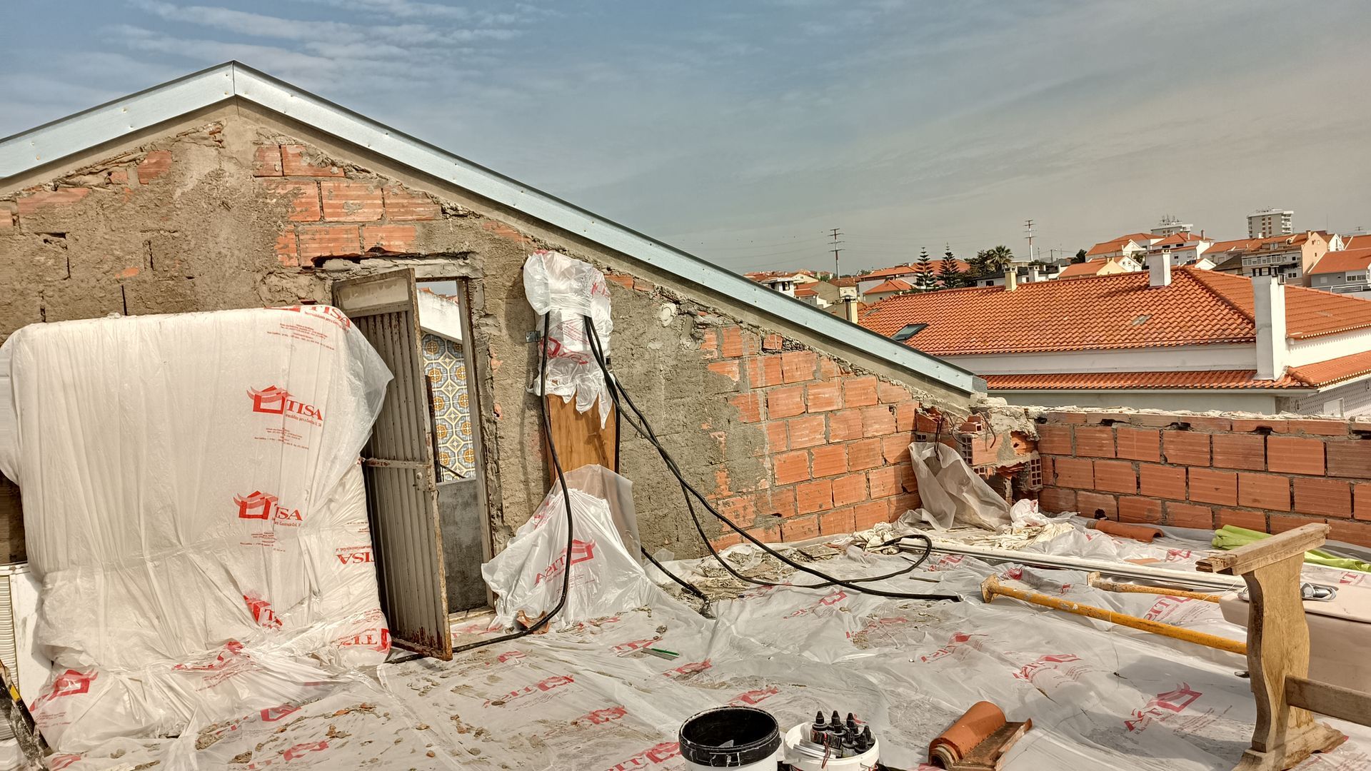 Construção no telhado: parede de tijolos inacabada, lona protetora, janela pequena e telhados de telha laranja ao longe.