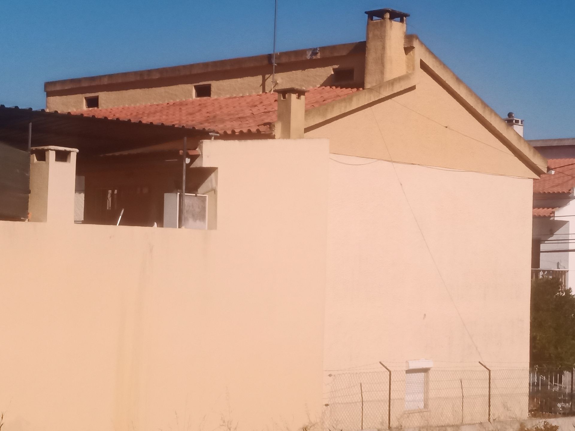 Prédio bege com telhado de telha vermelha, chaminé e área coberta; céu azul.