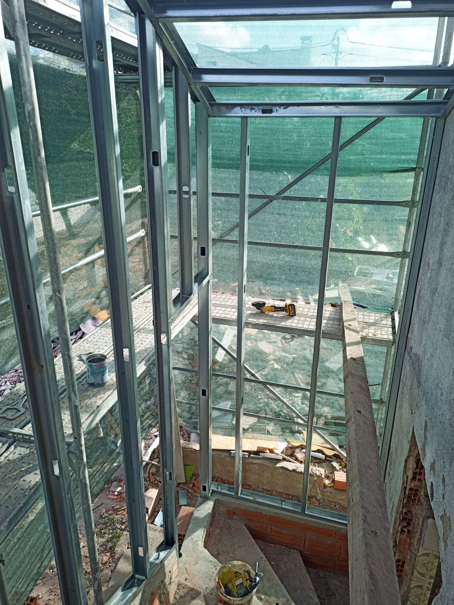 Estrutura metálica de um edifício em construção, escadas com rede verde e uma parede parcialmente acabada.