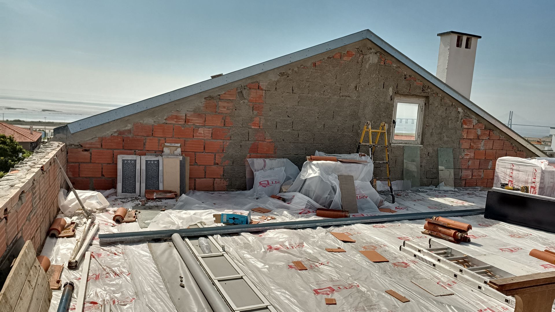 Canteiro de obras: telhado com tijolos aparentes, materiais de construção e chaminé em um dia ensolarado.