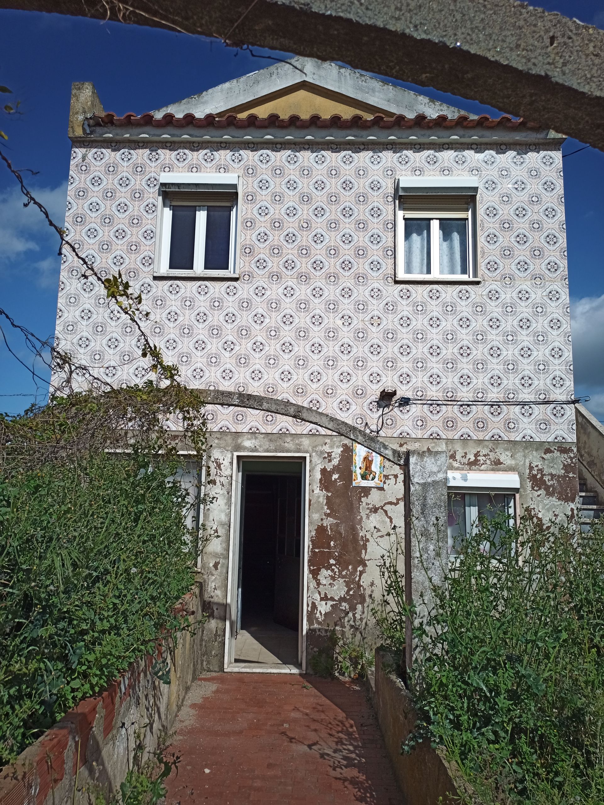 Prédio de dois andares com fachada de azulejos estampados, porta aberta e vegetação excessiva.