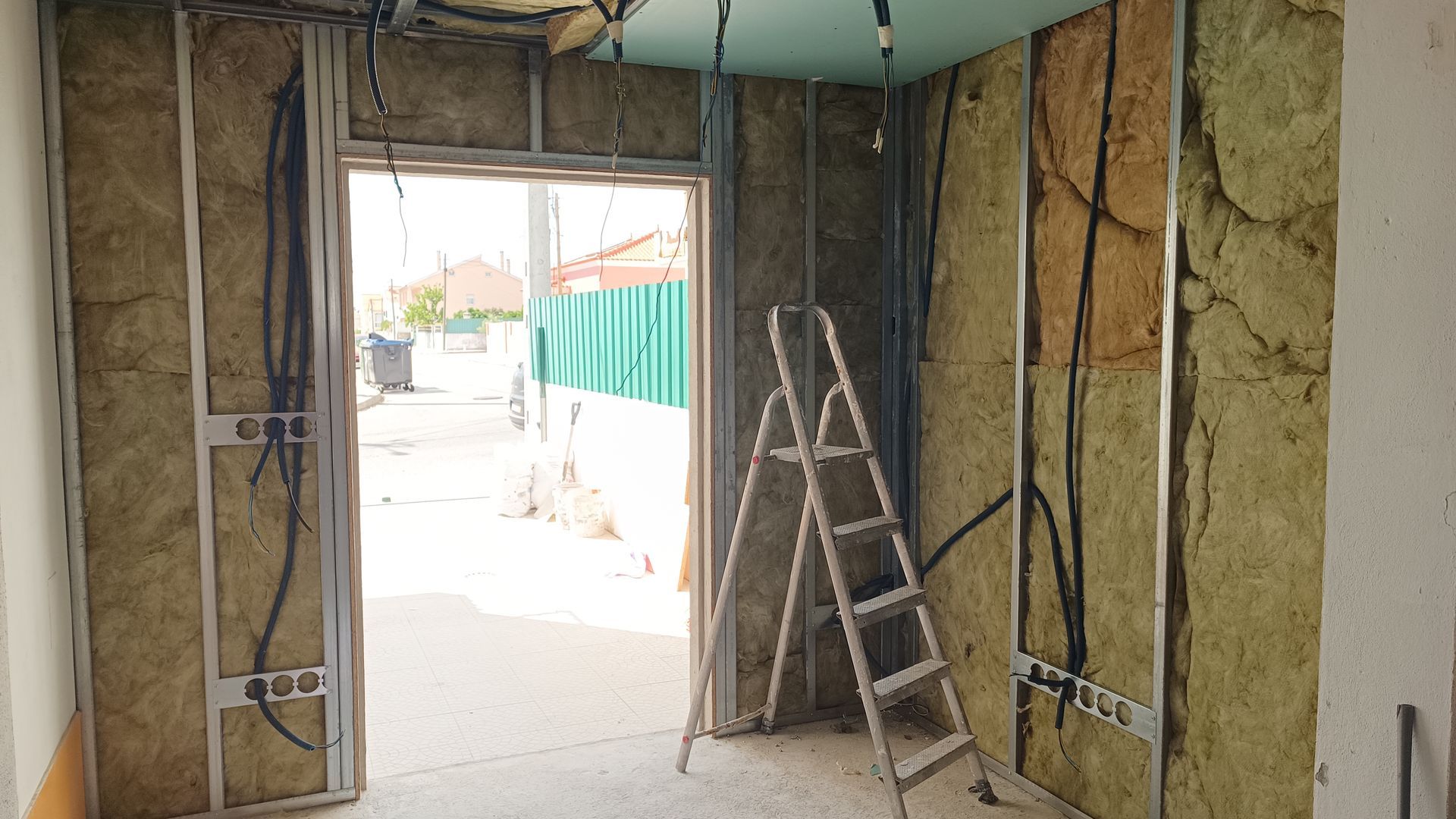 Construção de cômodo interno; isolamento exposto, estrutura metálica, escada, porta aberta para a rua.