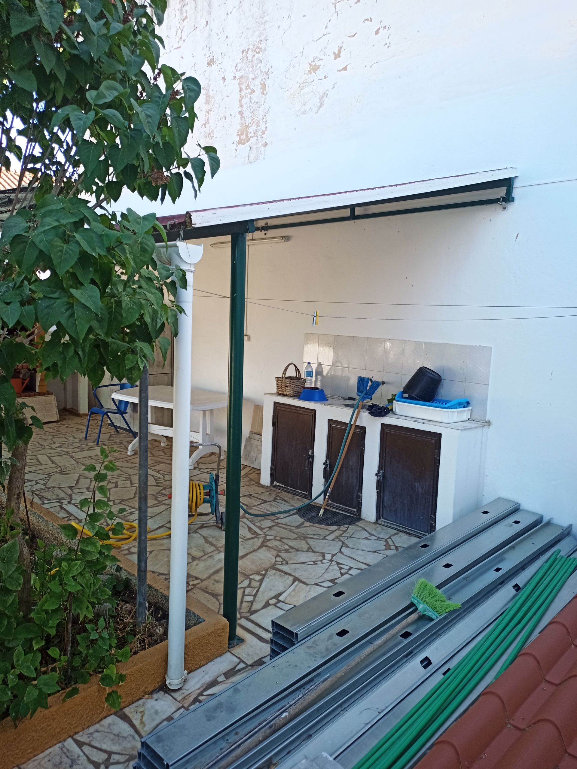 Cozinha externa sob um pátio coberto com armários brancos e piso de pedra. Pilares de sustentação verdes.